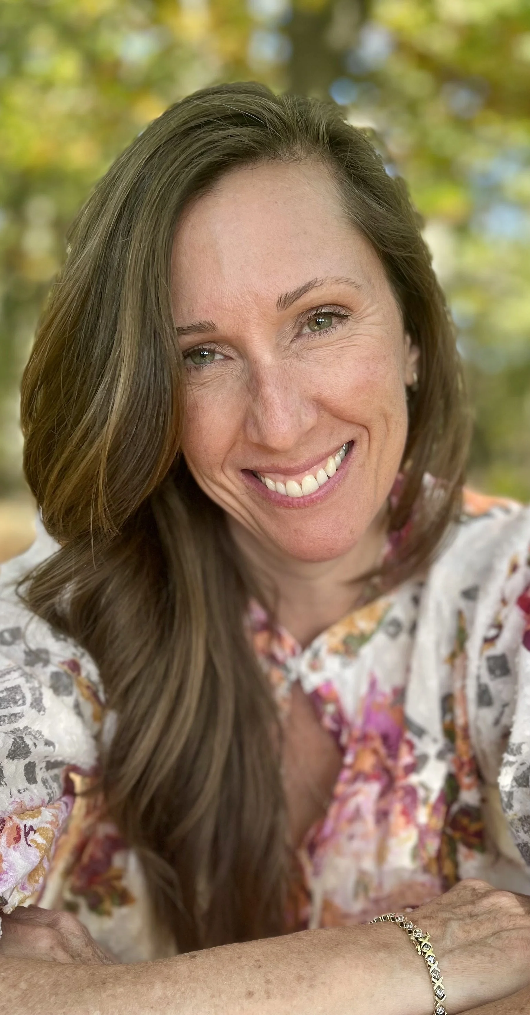 A smiling woman with long brown hair, wearing a floral blouse, outdoors with a blurred background of green and yellow trees.