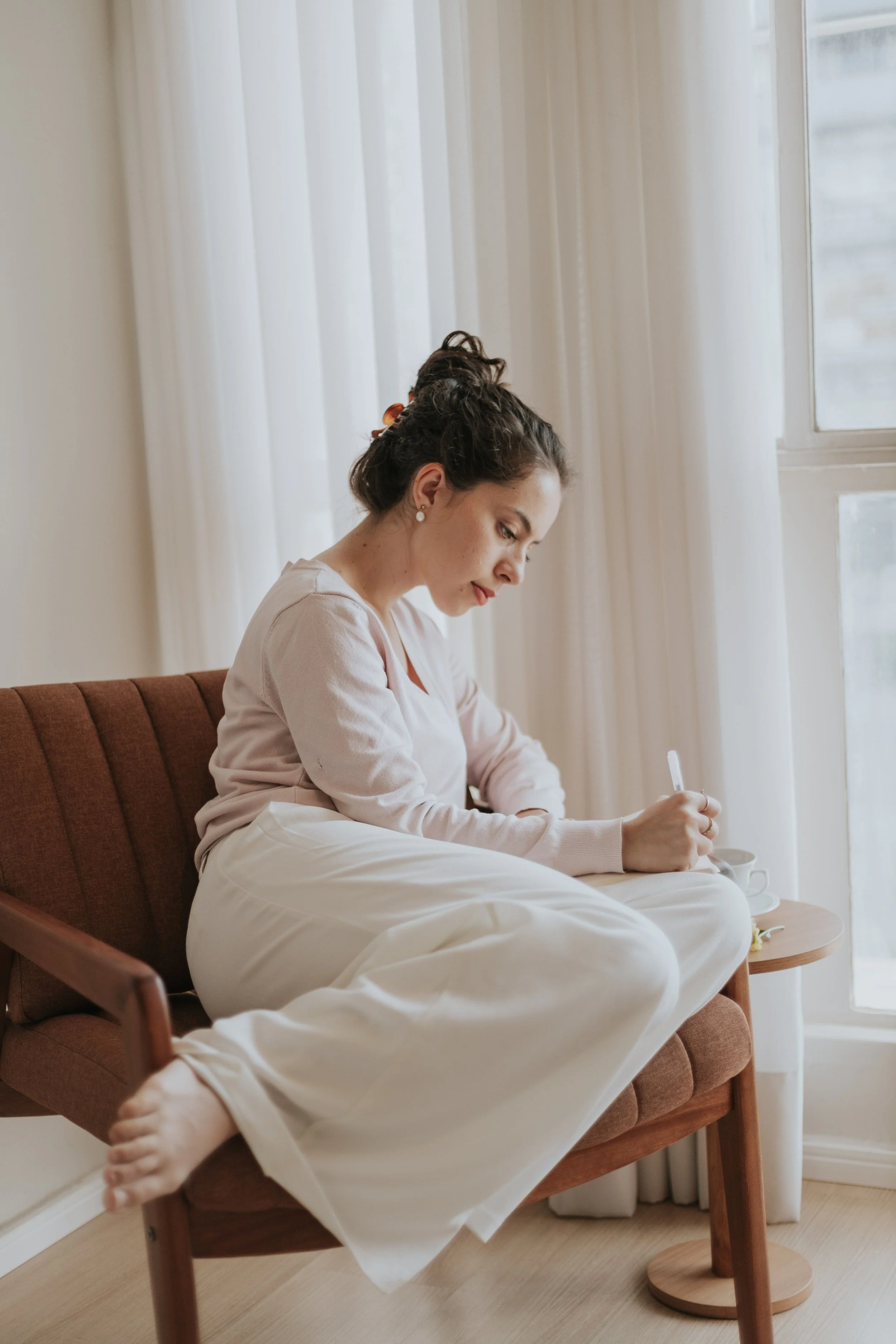 A woman sitting on a wooden chair with a cushioned brown seat, writing in a notebook by a large window with white curtains, wearing a light-colored long-sleeve shirt and white pants, with a cup of coffee or tea on a small round side table.