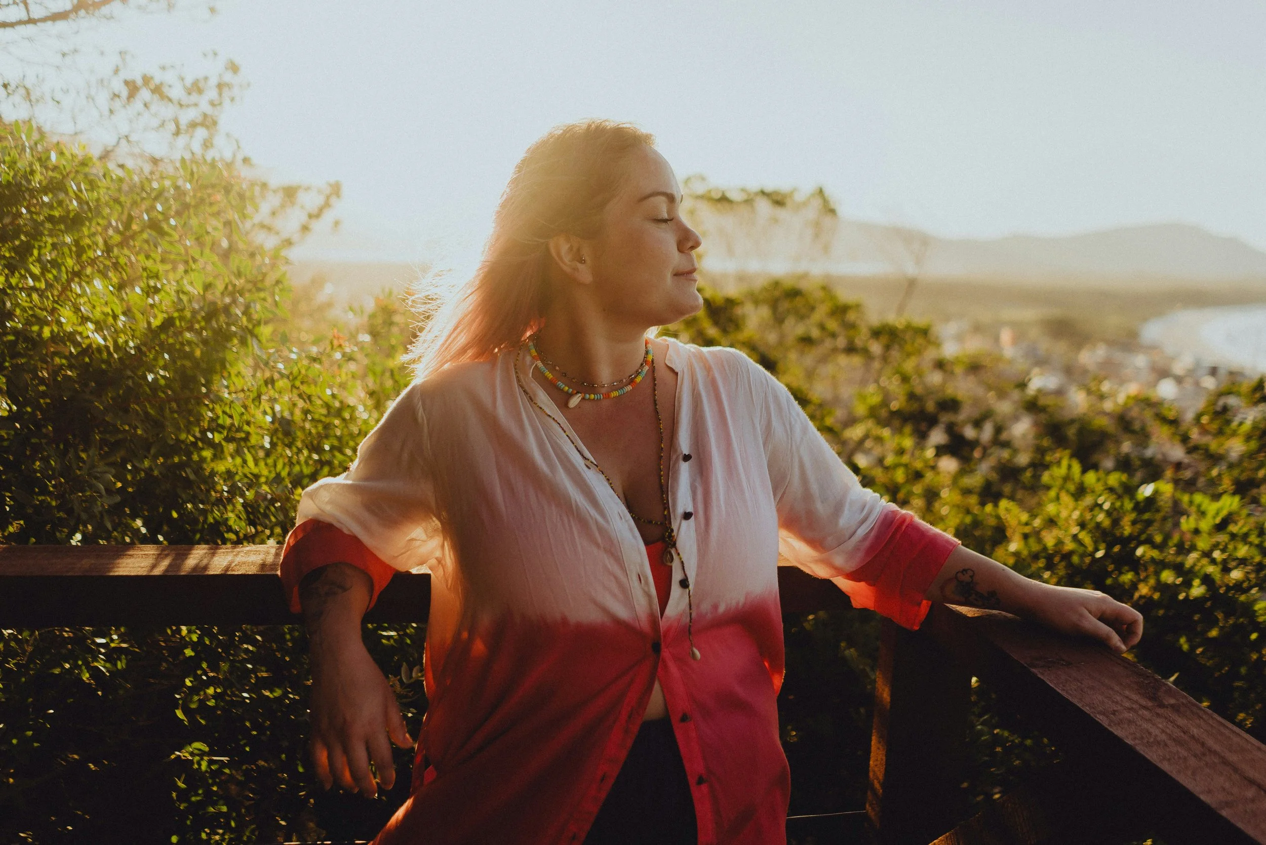 Woman with closed eyes and colorful jewelry resting on a wooden railing during sunset.