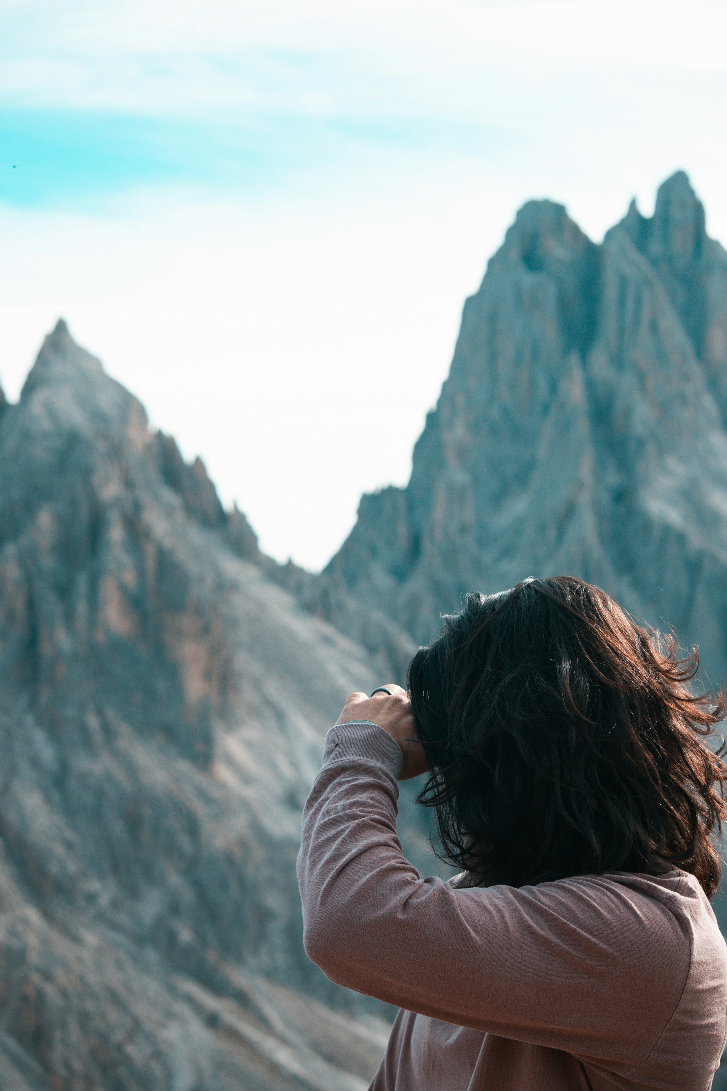 A person with dark hair taking a photo of mountainous landscape with rugged peaks, wearing a light gray or beige jacket.