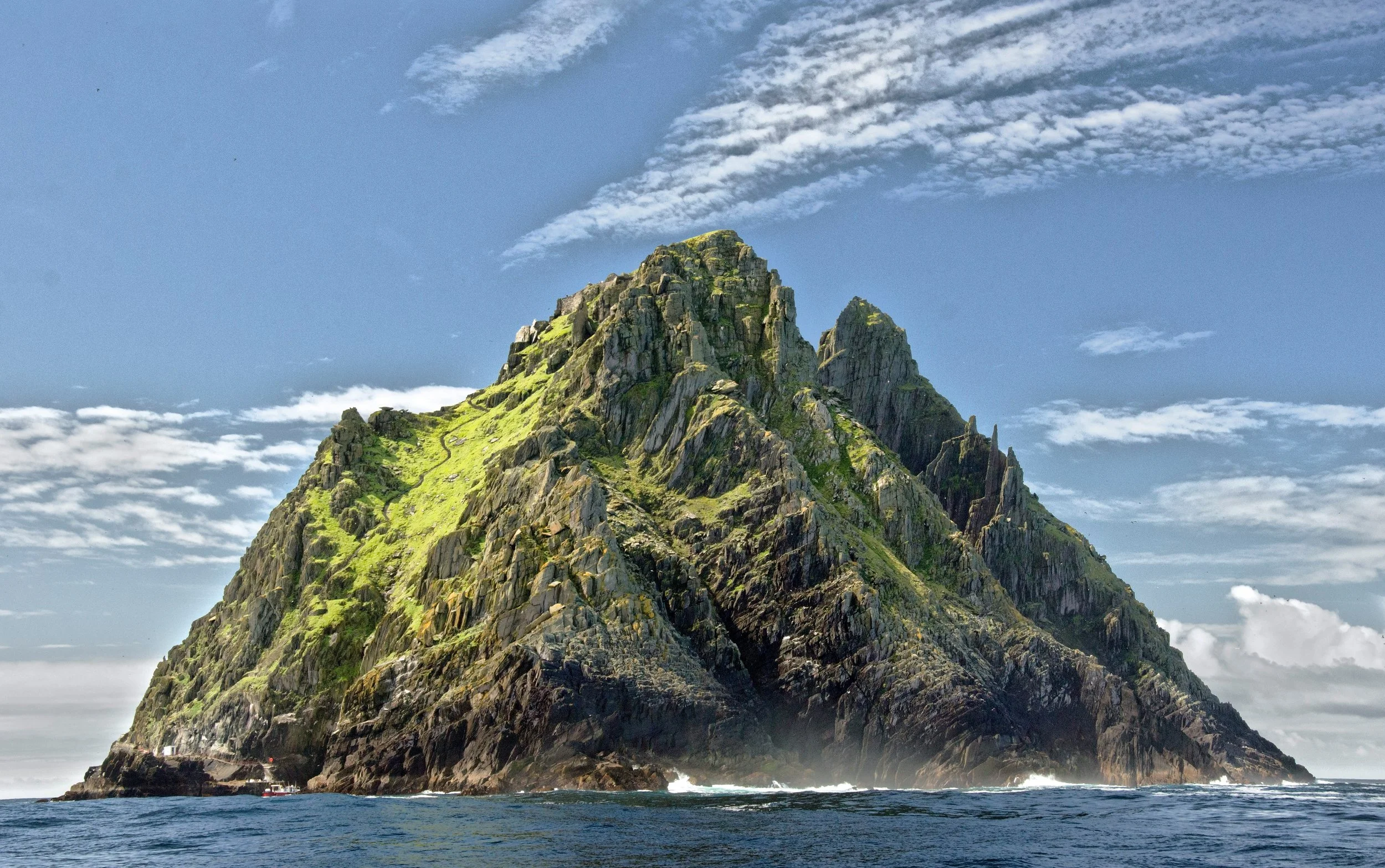 A tall, rocky, green-covered mountain rising from the ocean, with a partly cloudy sky in the background and boats at the base.