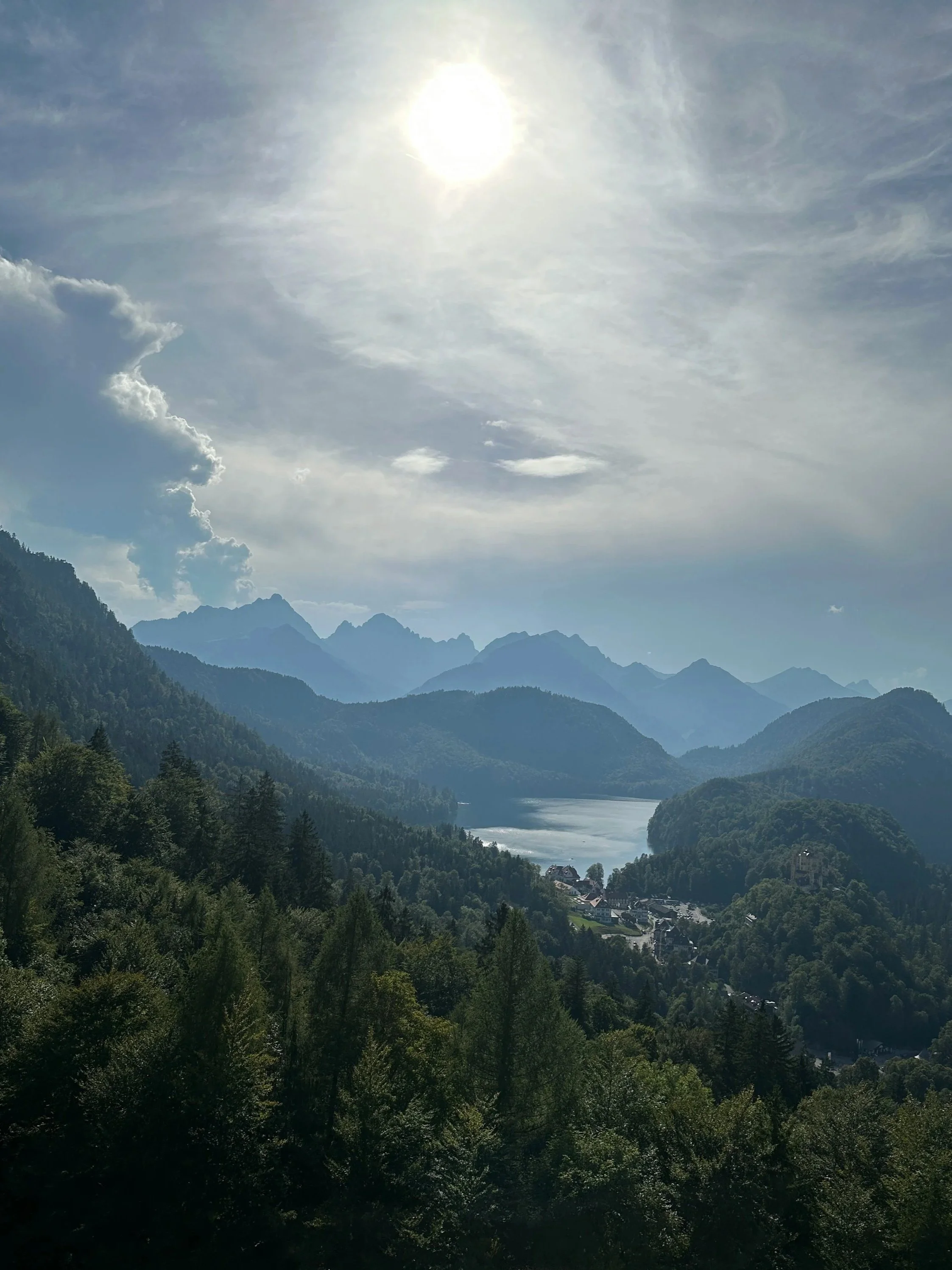 Scenic view of a river flowing through a lush green valley surrounded by tall mountains under a bright sun and partly cloudy sky.