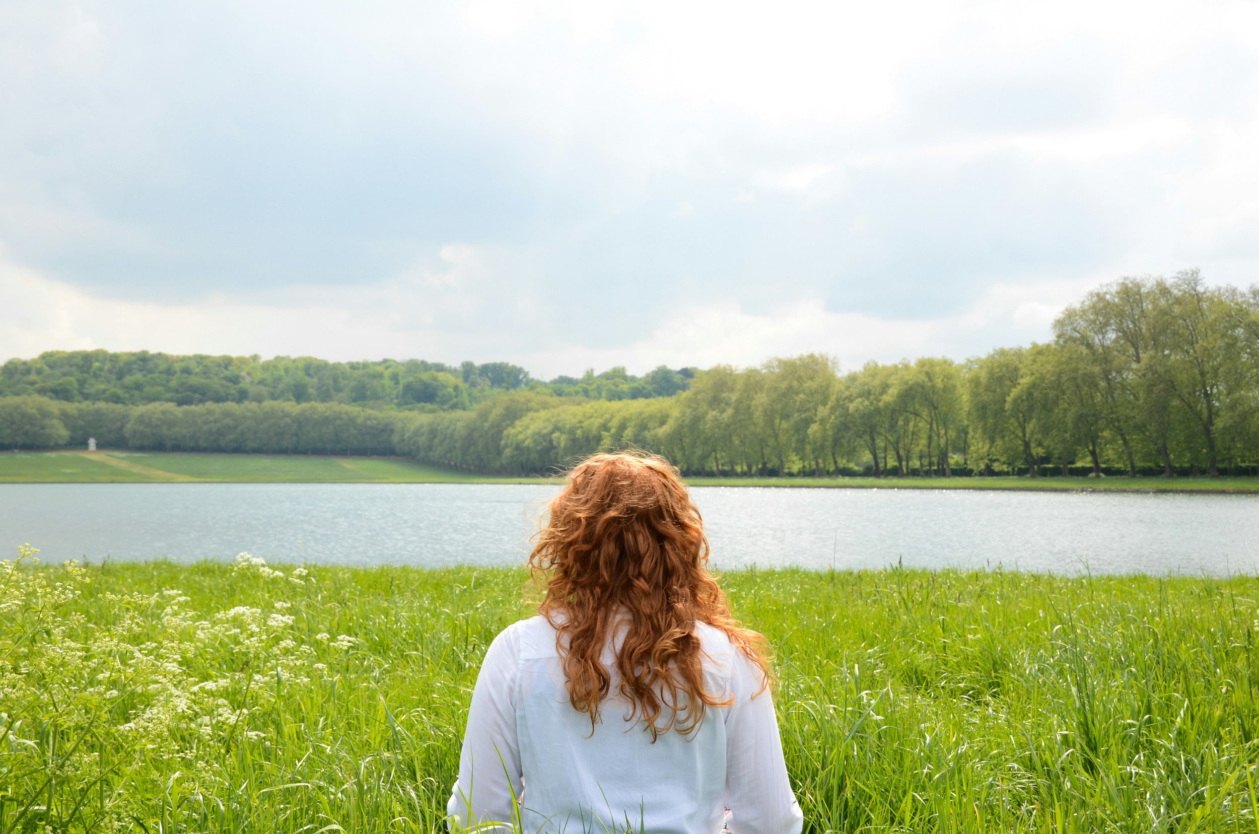 Person with long red hair sitting on grass by a lake, facing away, with green trees and cloudy sky in the background.