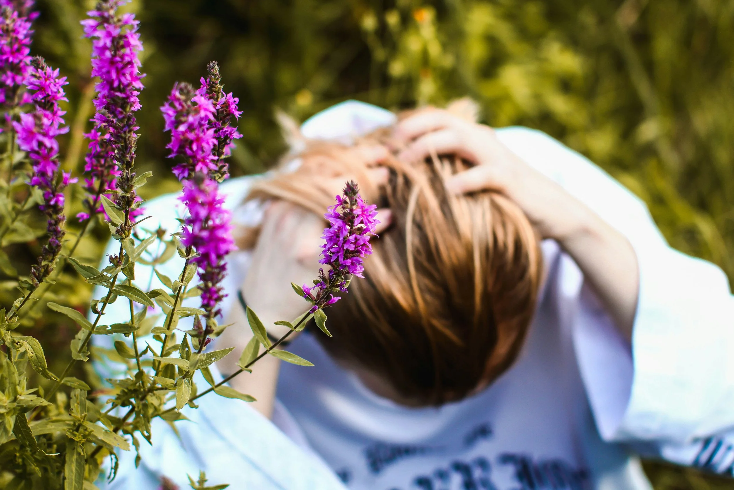 A woman with long light brown hair, wearing a white shirt, is outdoors with head in hands in sunlight near purple flowering plants.