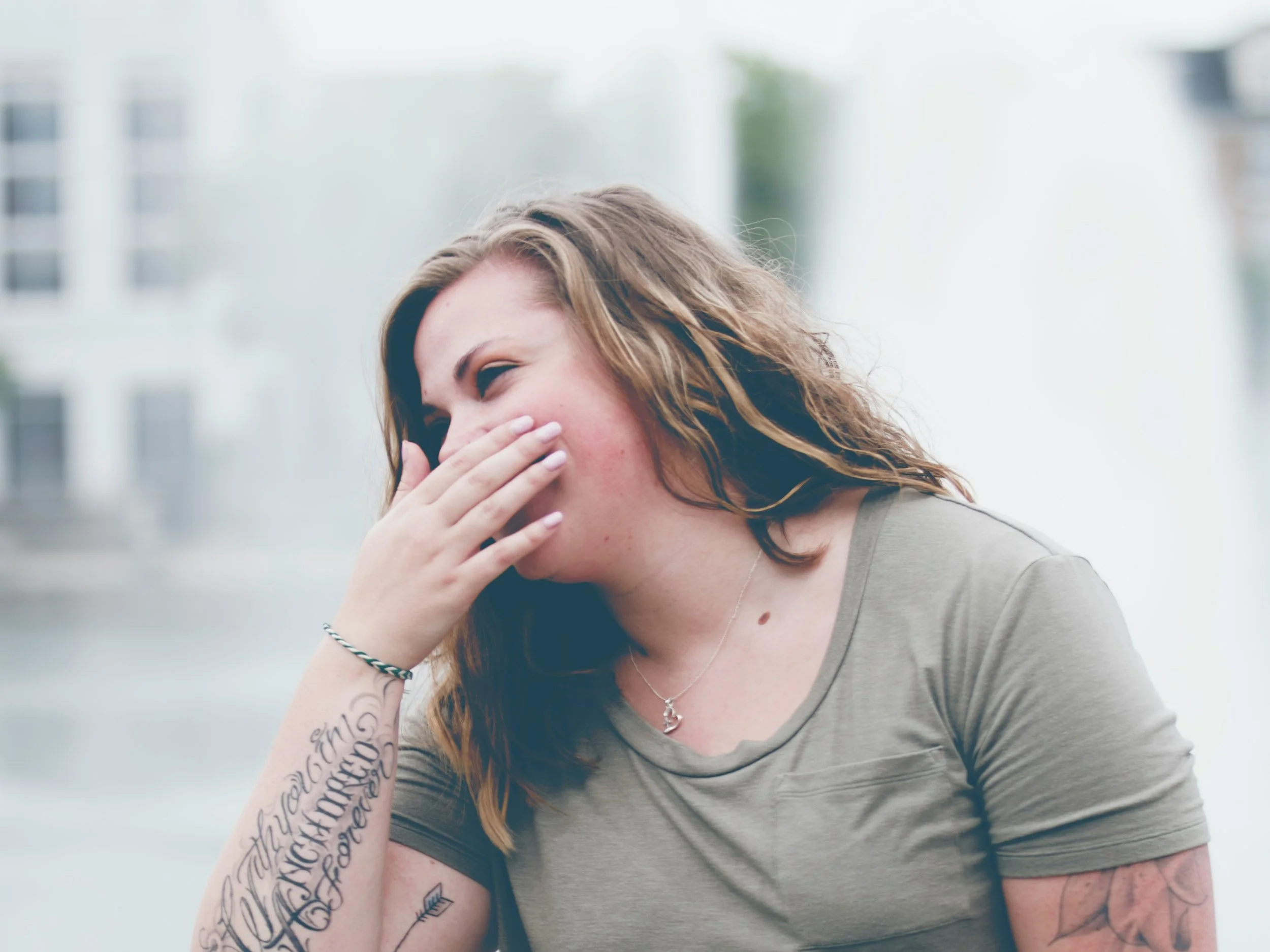 A person with brown hair covering her face, hand over her mouth, showing tattoos on her arm and a necklace with a pendant, standing outdoors with a blurred cityscape background.