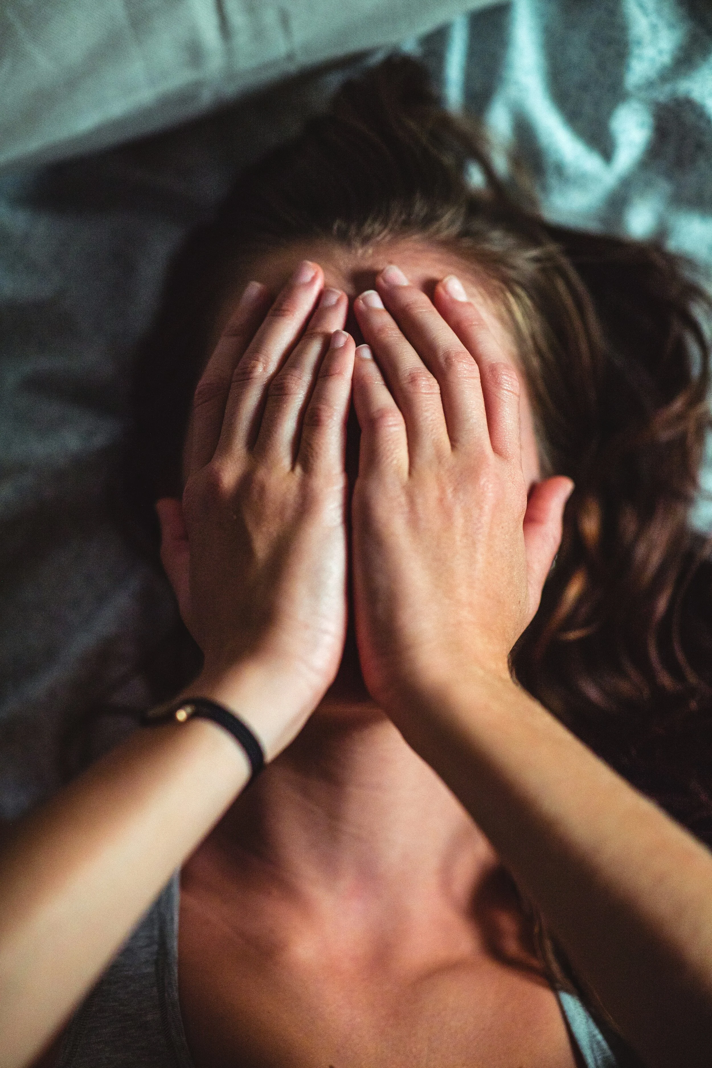 Person with brown hair covering their face with hands, lying down with another person's hands on their face, surrounded by gray fabric.