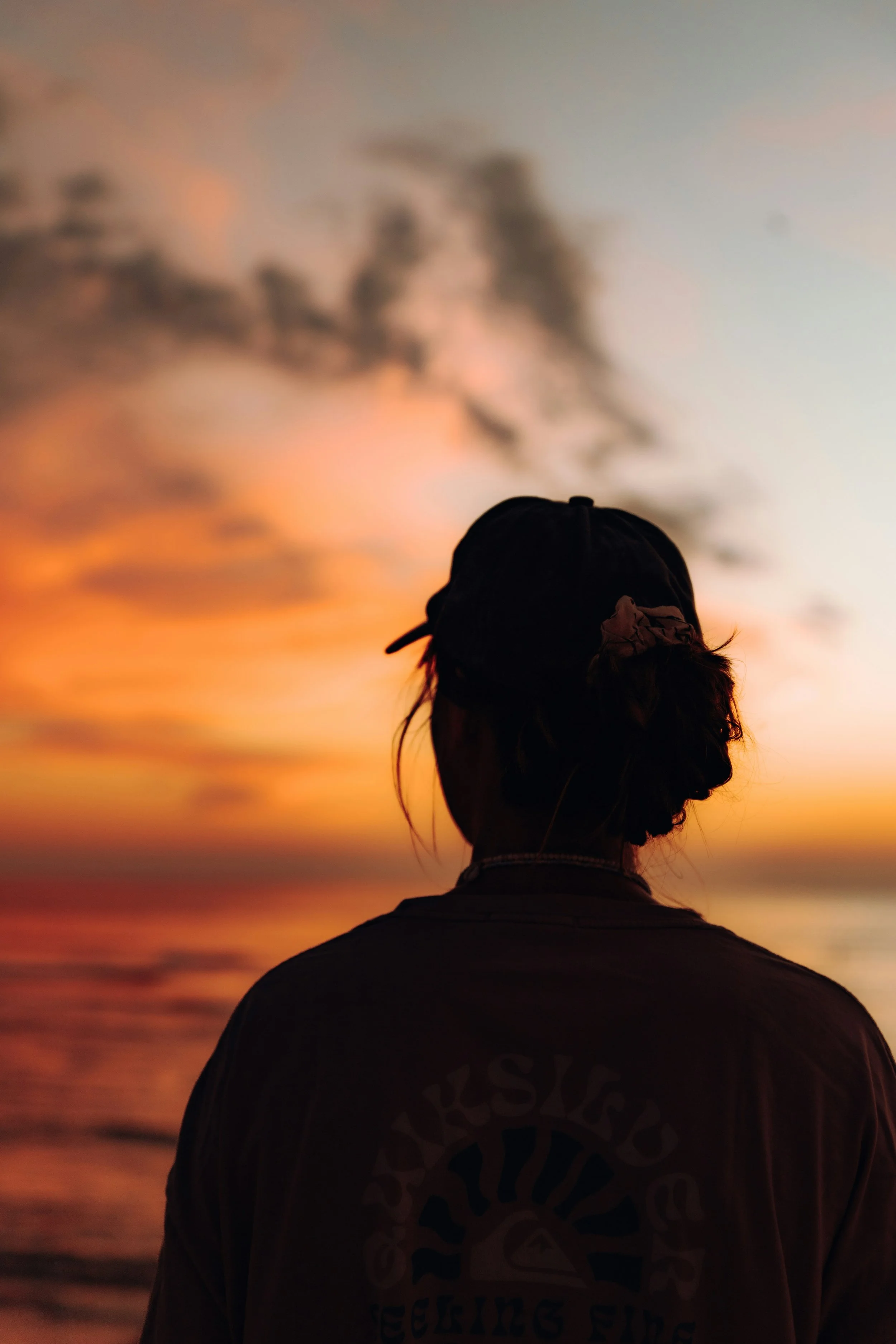 A woman standing at the beach, calmly watching a sunset sky painted in shades of orange, pink, and blue. The person's face is not visible.