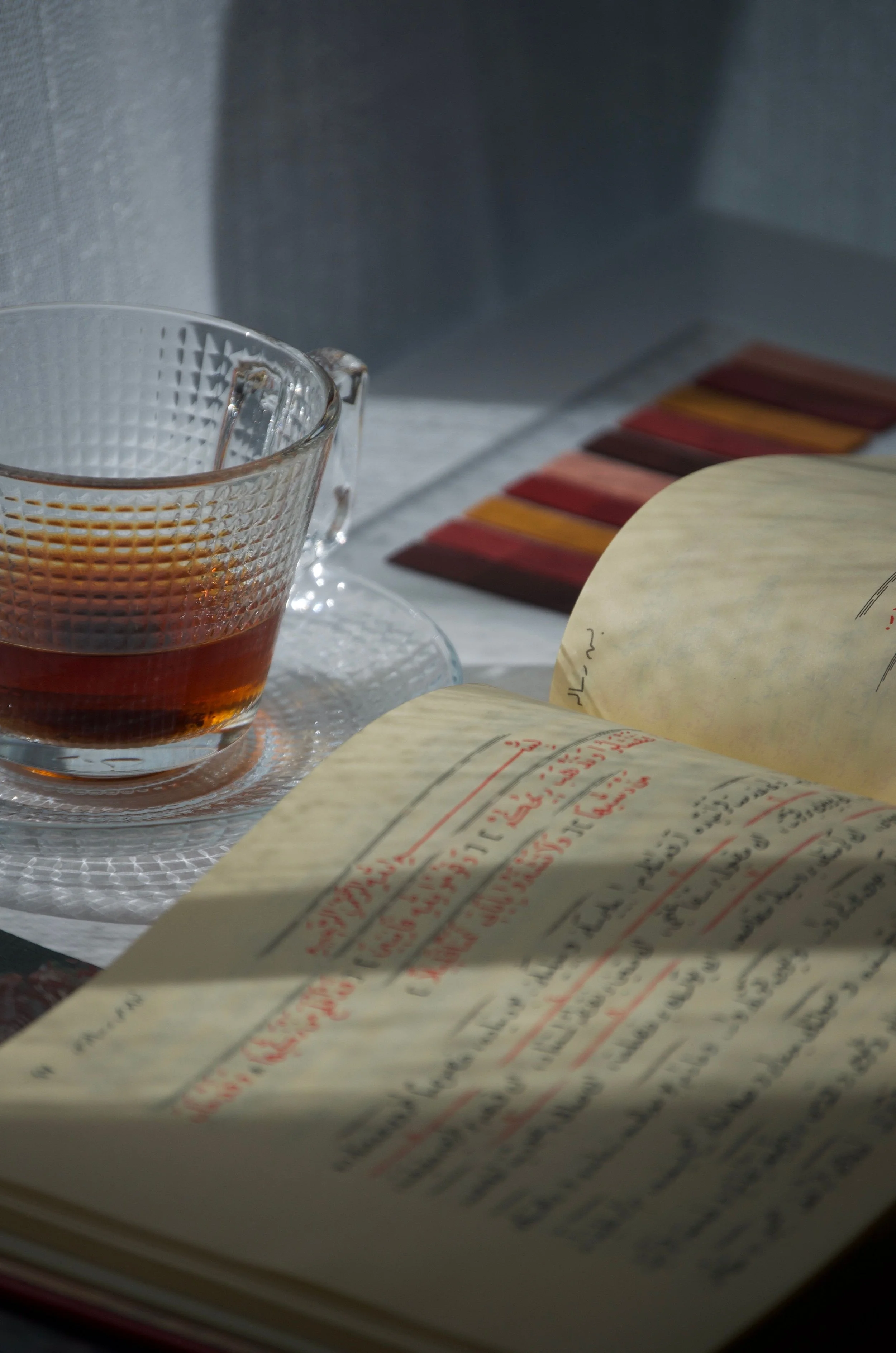Open book with red and black handwritten text on cream-colored pages, a glass cup with brown liquid, and a colorful striped item in the background, all on a white surface.