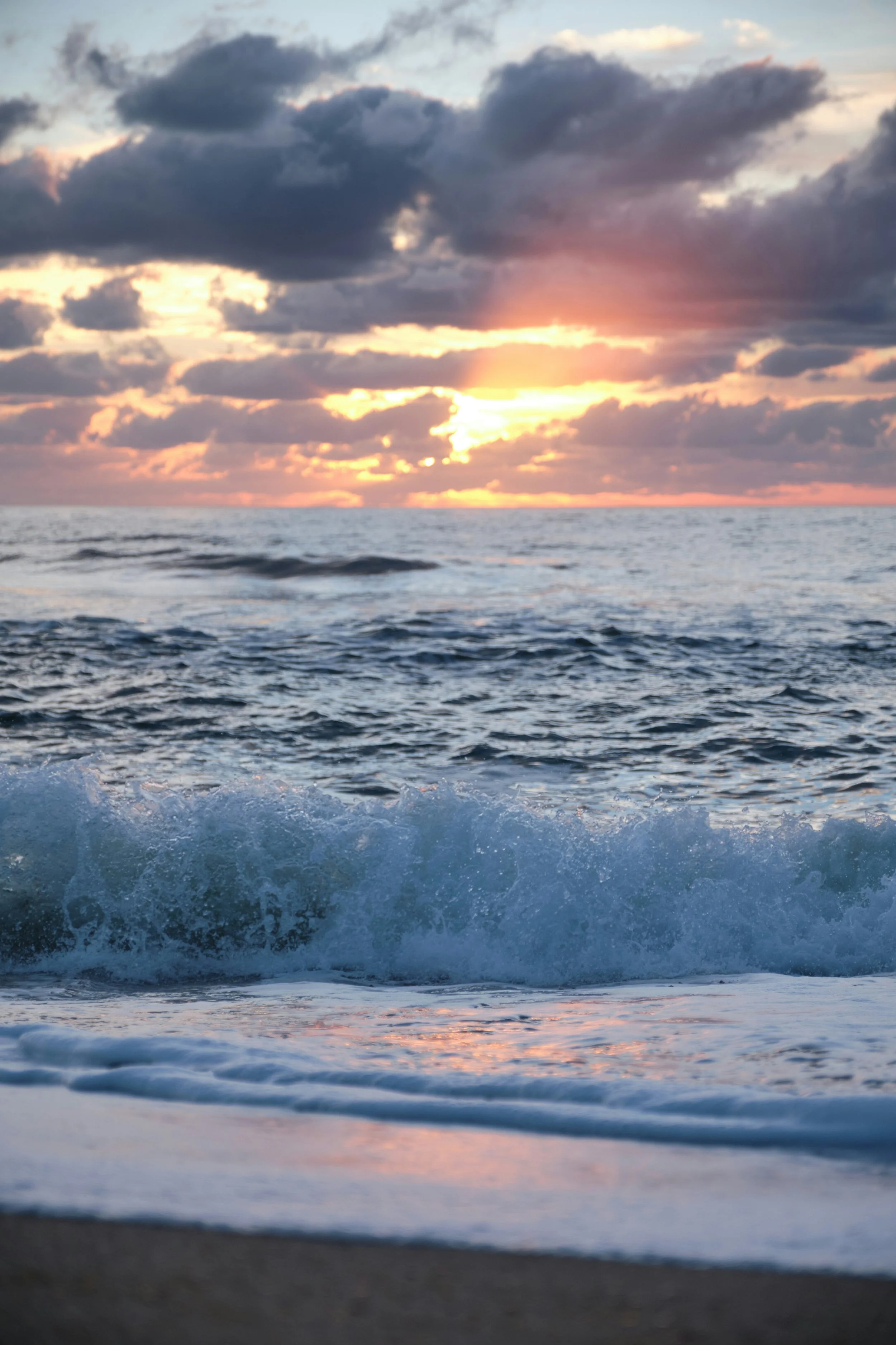 Sunset over the ocean with cloudy sky and waves crashing on the shore.