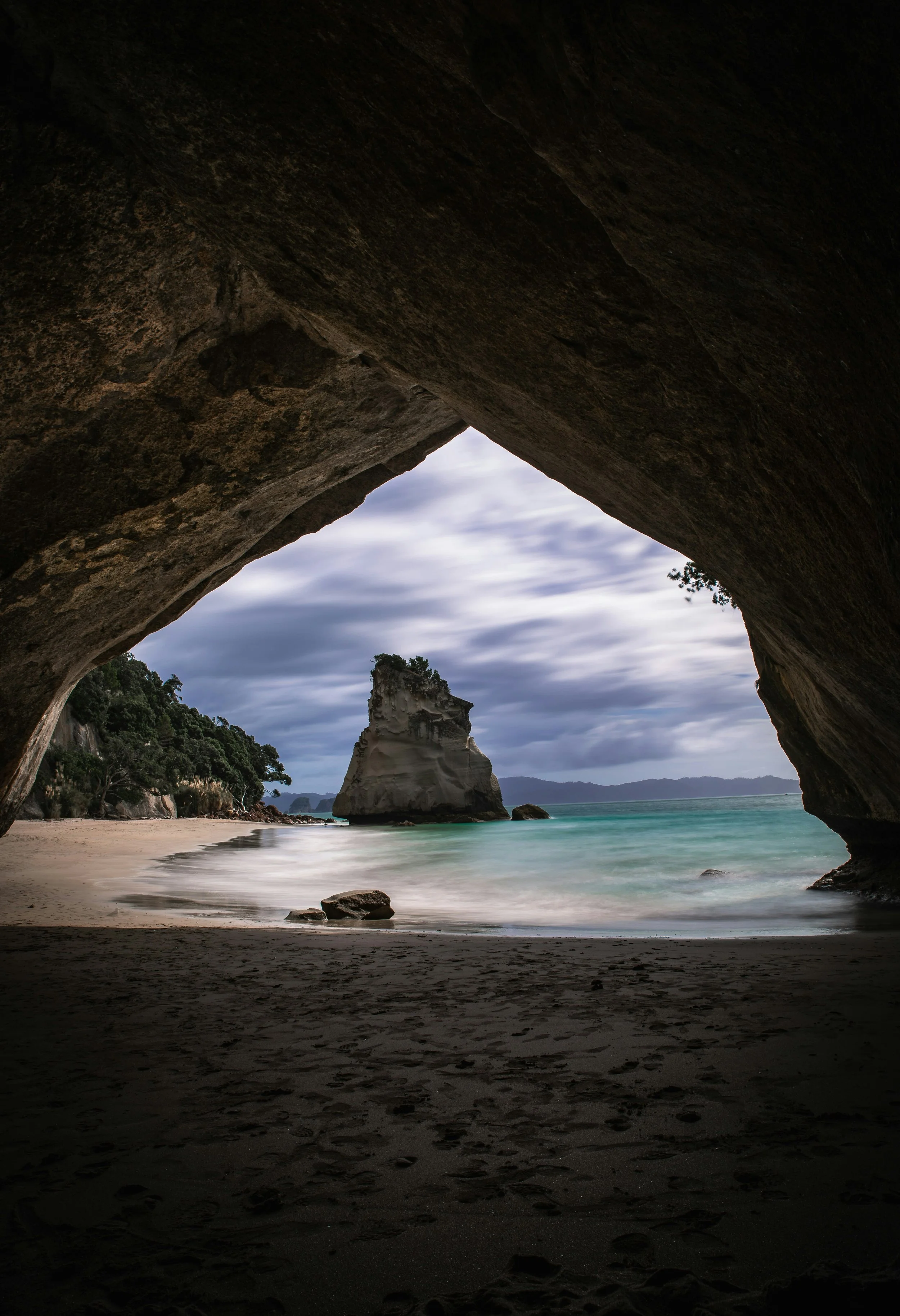 A view of a beach cave opening with dark shadows in the foreground, a sandy shore, rocky formations, and a distant white rock formation in the water under a cloudy sky.