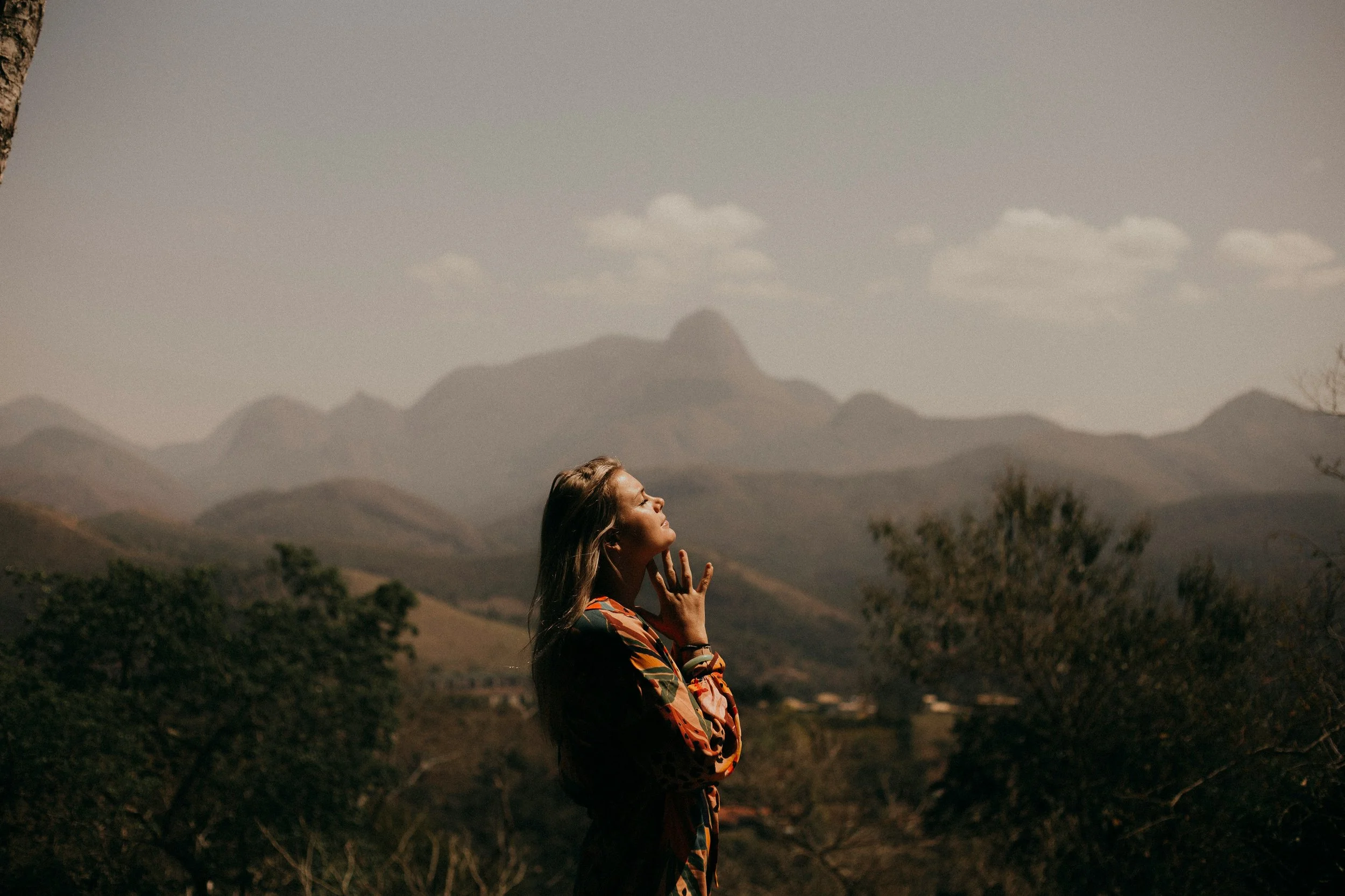 A woman with long hair standing outdoors in front of a mountain range, looking upward with her hand near her chin, in a natural landscape setting.