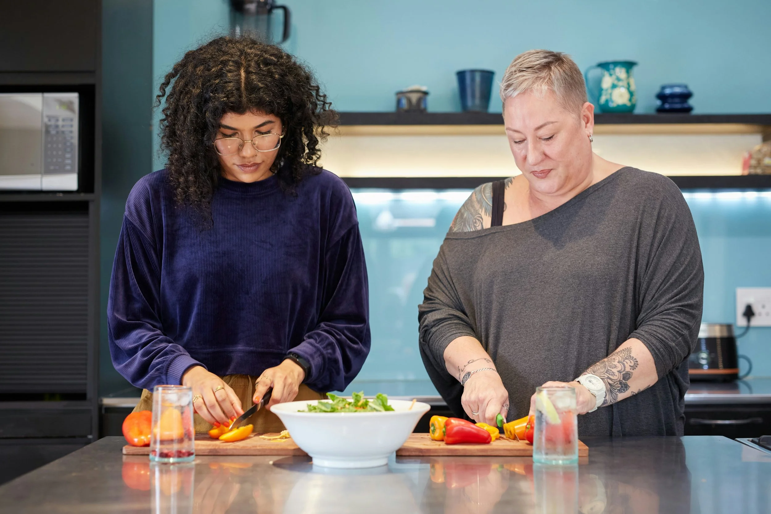 Two women preparing vegetables for a salad in a modern kitchen. One woman has curly hair, glasses, and is wearing a purple velvet shirt; the other has short blond hair, tattoos on her arm, and is wearing a gray top. There are bell peppers, tomatoes, and a salad bowl on the counter.