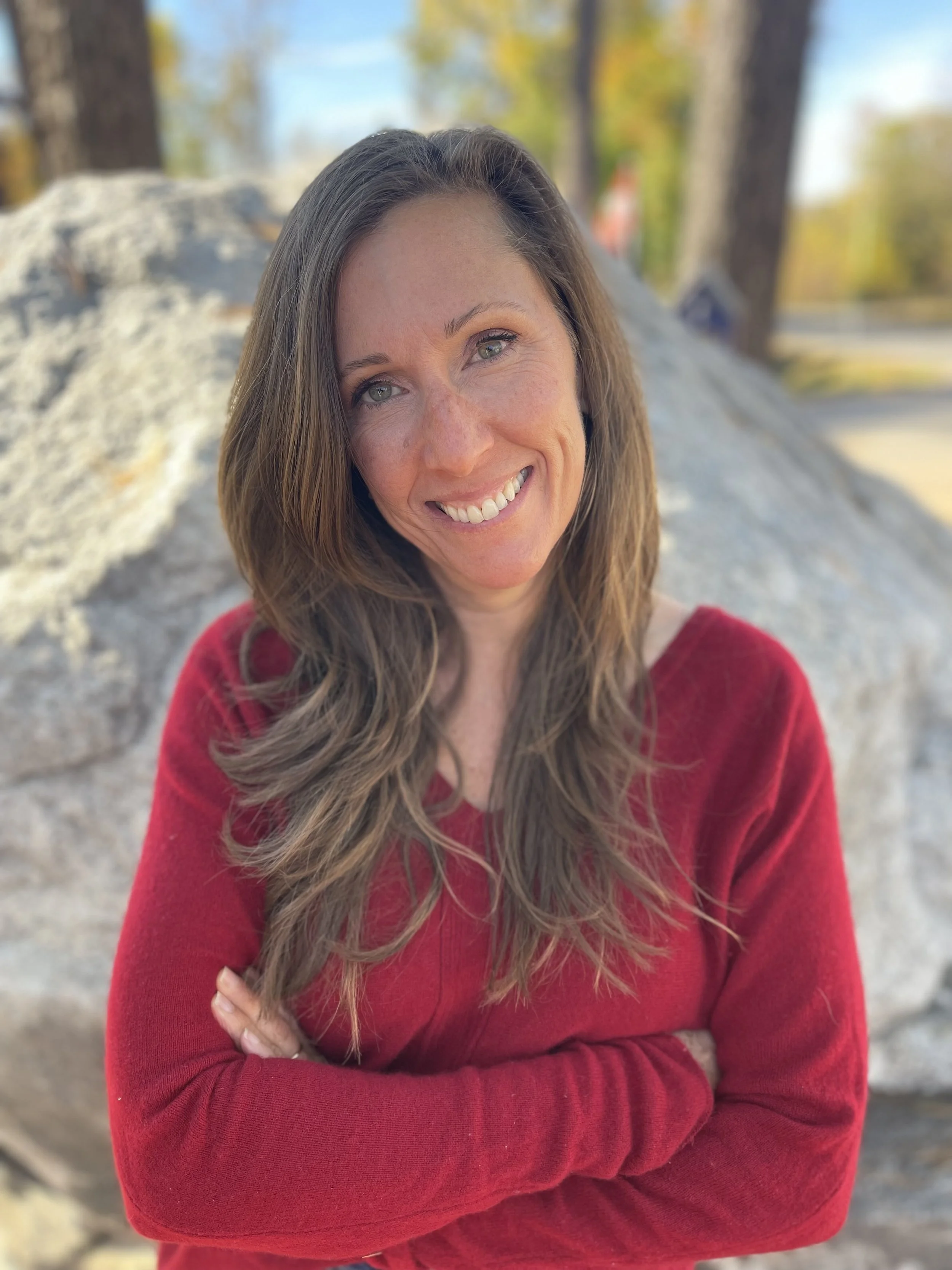 A woman with long wavy brown hair and blue eyes smiling, wearing a red sweater, standing outdoors in front of rocks and trees with autumn-colored leaves.
