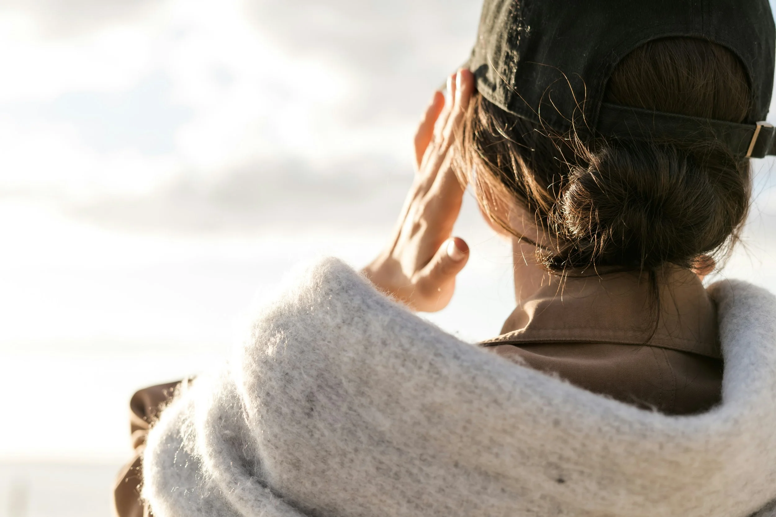 Person wearing a black beanie and beige coat, holding their hand to their face, outdoors in snowy weather with sunlight.
