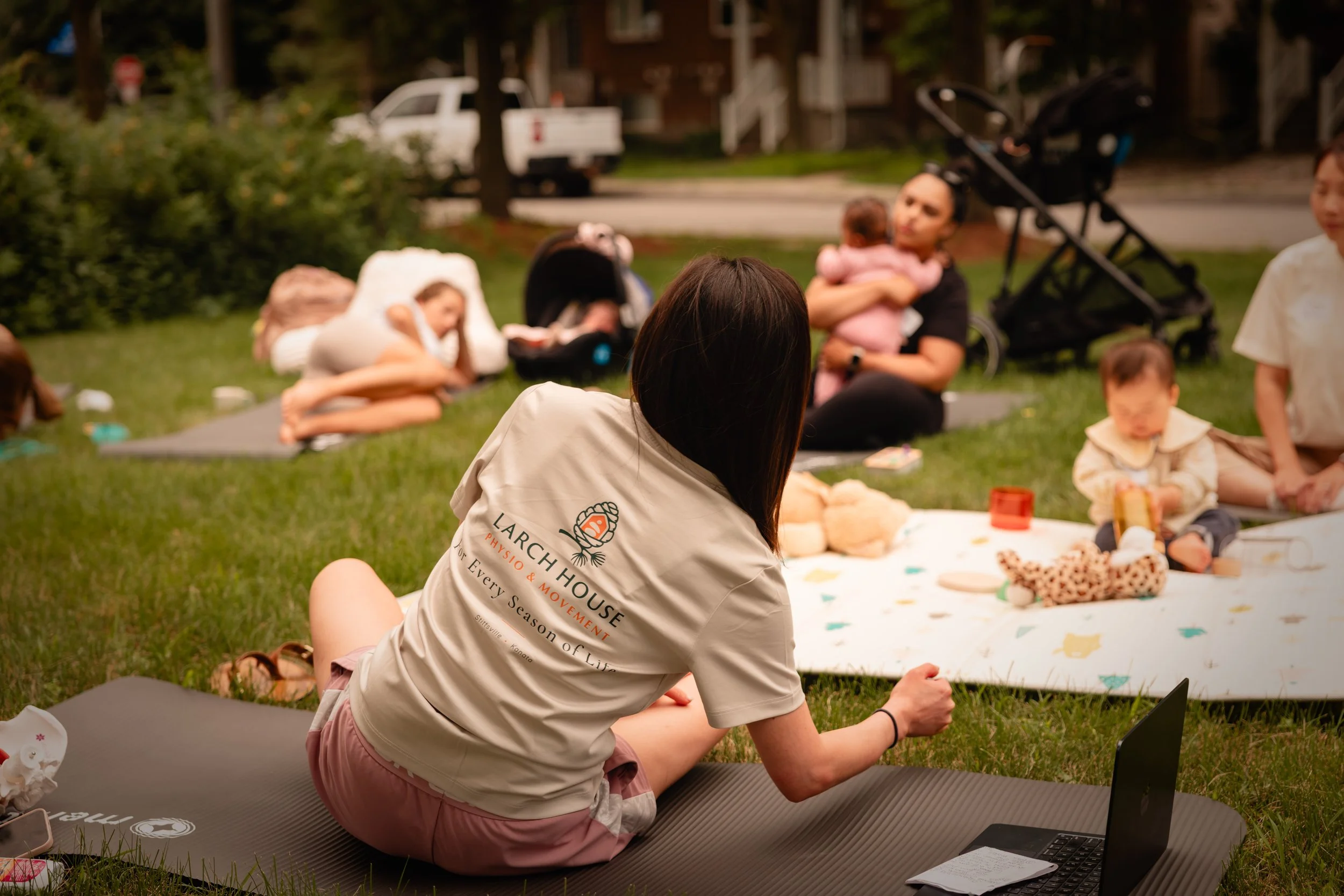 A woman sitting cross-legged on a yoga mat outdoors, wearing a beige t-shirt and pink shorts. She is participating in a virtual meeting or class on her laptop, with children and other people in the background, some sitting on the grass, and a stroller visible. The scene appears to be a gathering in a park or backyard.