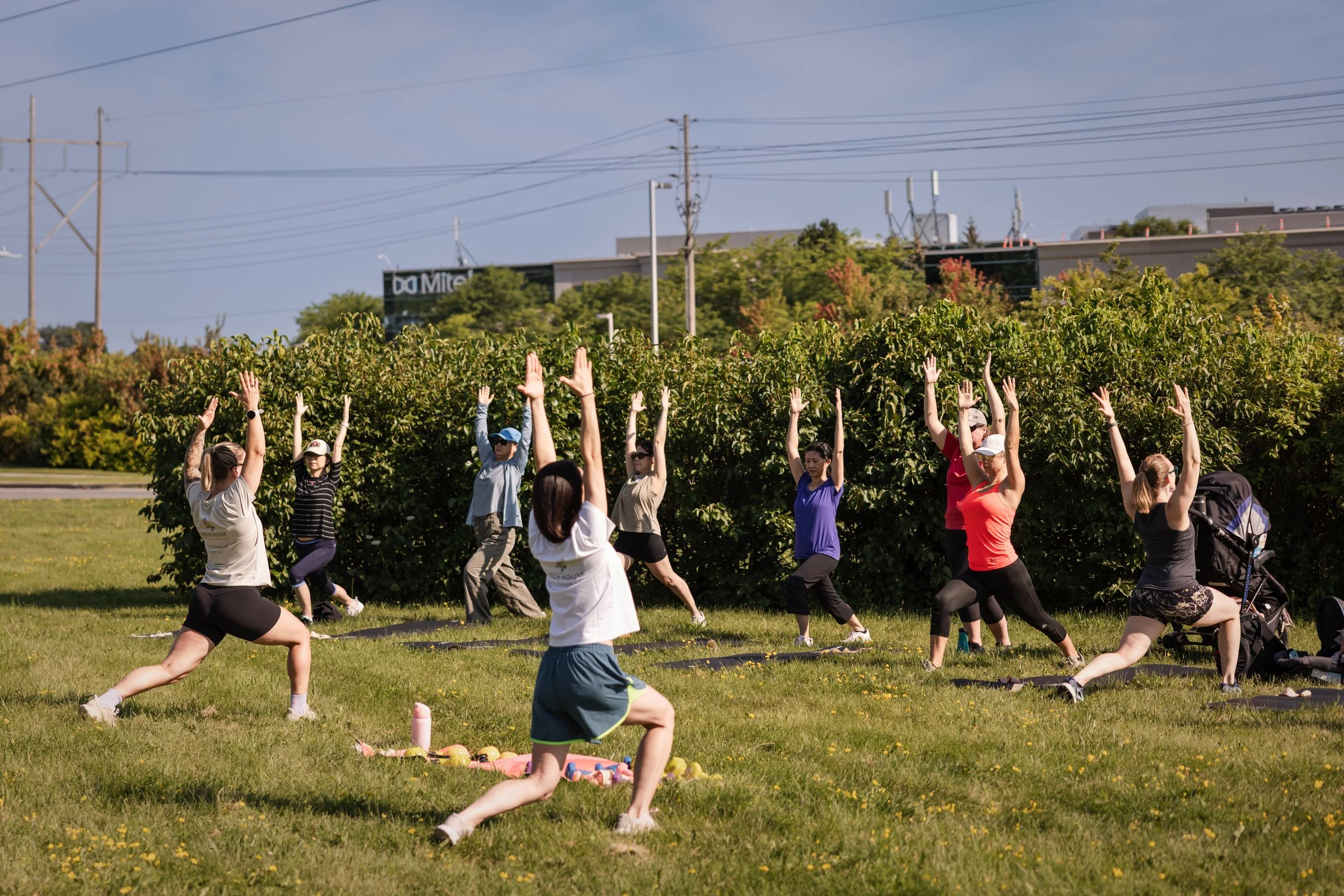 Group of people participating in an outdoor yoga or fitness class on grass, raising their arms in a pose, with trees and utility poles in the background.