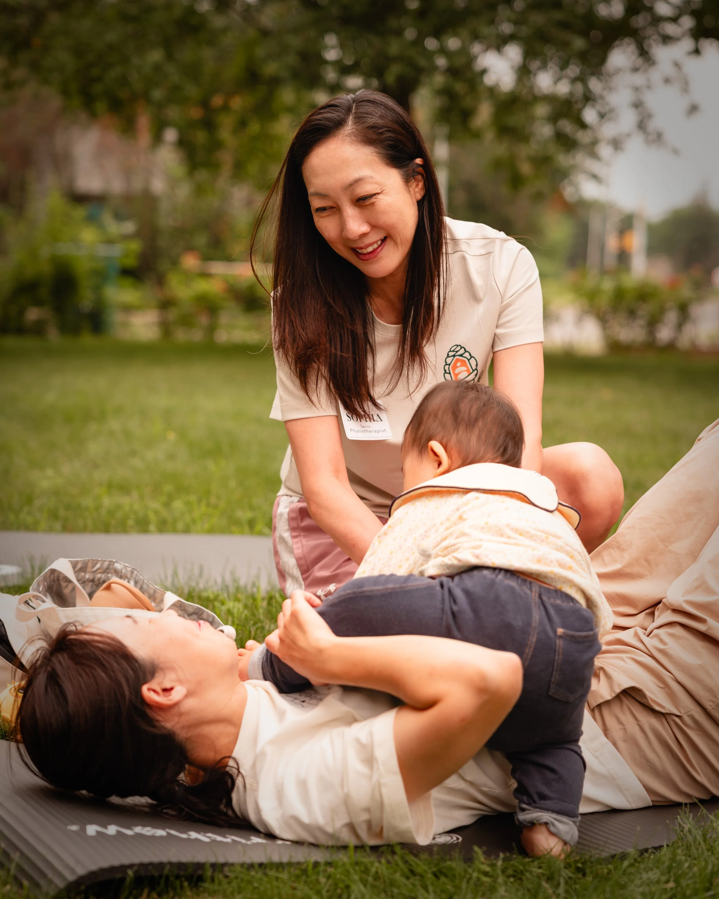 A woman practicing yoga outdoors on a mat in a park with children around her, one of whom is lying on her back.
