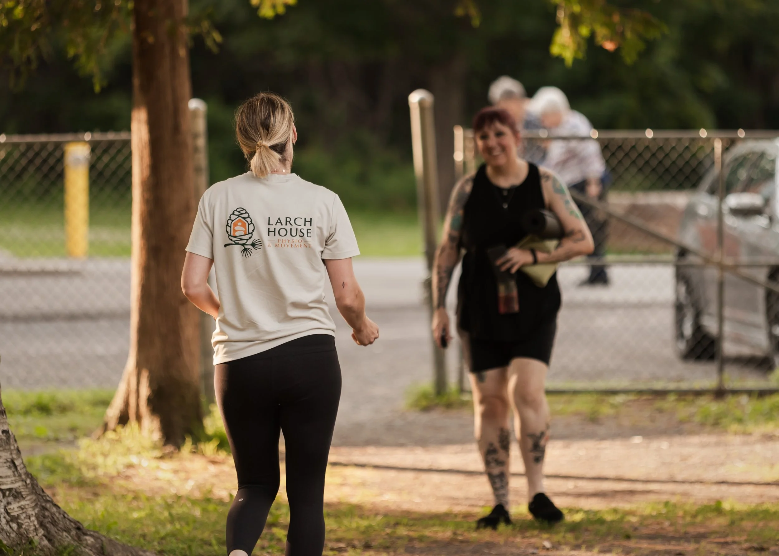 A woman with blond hair in a ponytail wearing a white t-shirt with the logo 'Larch House Physio & Movement' on the back, standing outdoors, facing another woman with tattoos, short red hair, and a black sleeveless top, in a park area near a chain-link fence and trees, during what appears to be an outdoor fitness or wellness session in the evening.