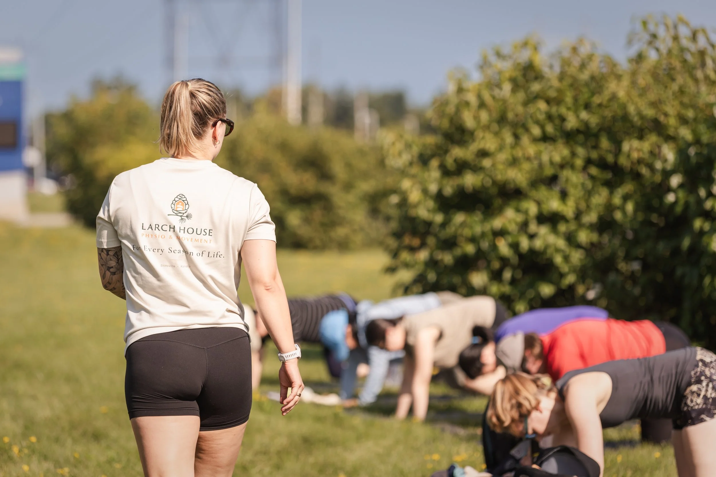 A woman in athletic attire leading an outdoor yoga or stretching class with people in a plank position on the grass.