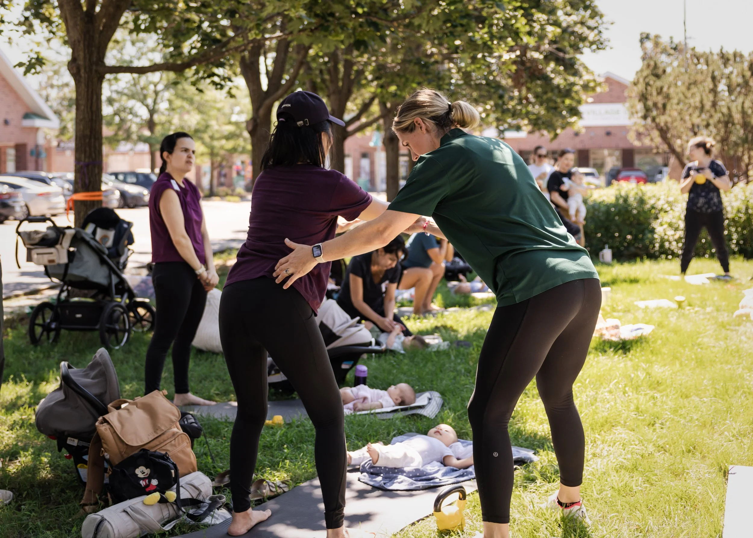 Two women practicing yoga outdoors on a sunny day, with a group of people and a baby in the background, surrounded by trees in a park.