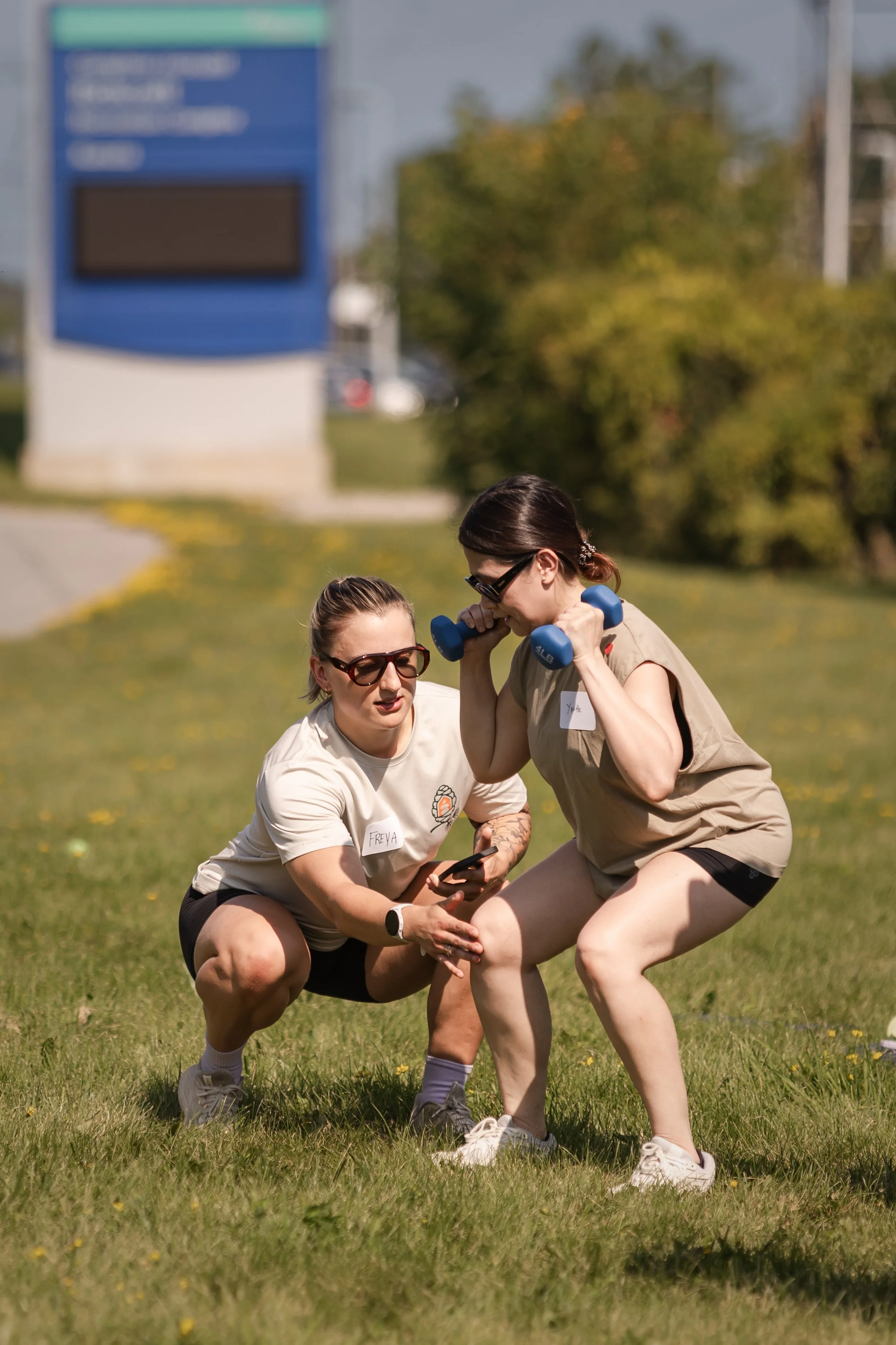 Two women in athletic clothing participating in an outdoor activity on a grassy field, one holding a dumbbell to her shoulder while the other appears to be assisting or coaching.