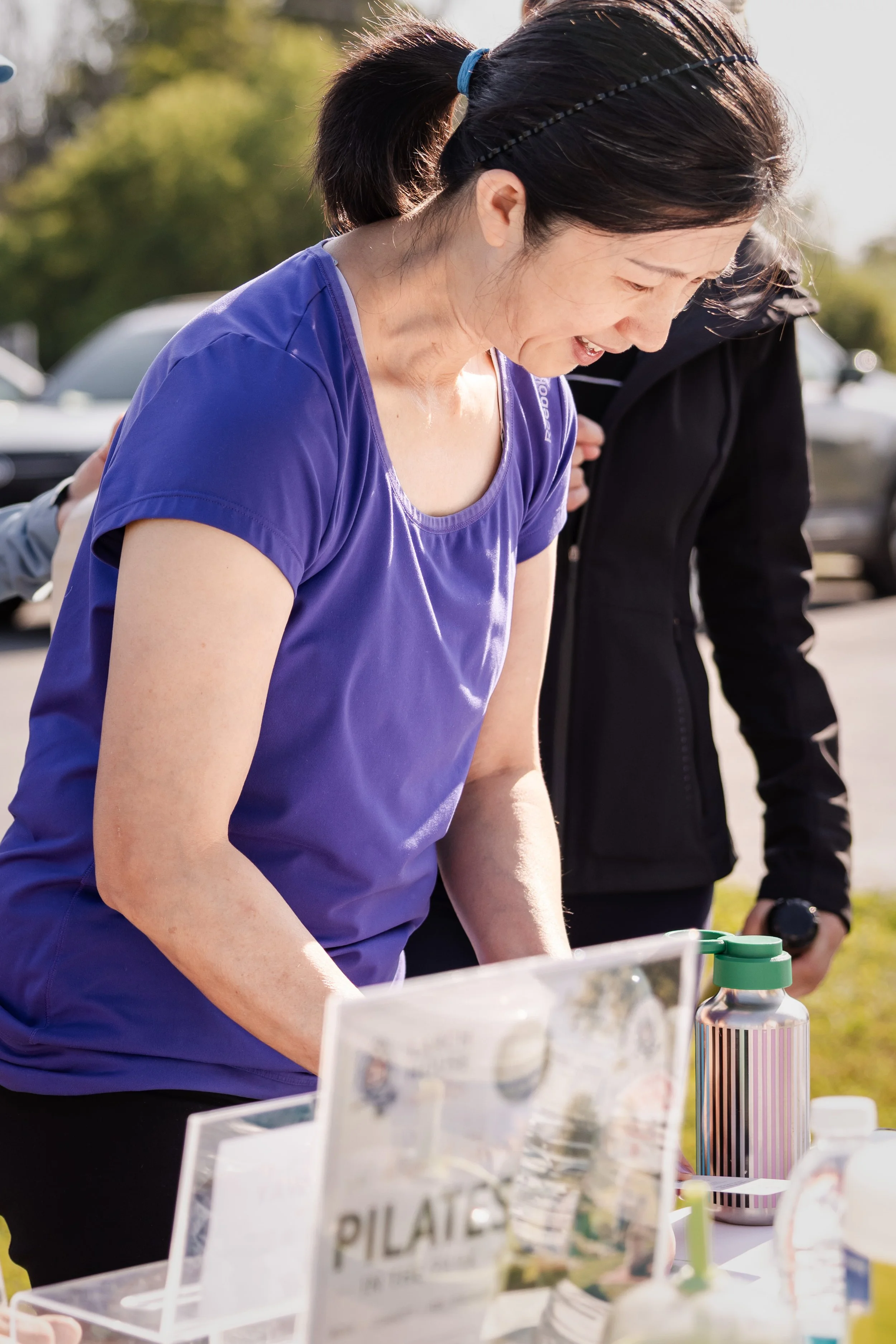 A woman in a purple shirt leaning over a table outdoors with various items, including water bottles and informational signs, with another person next to her. Trees and parked cars are visible in the background, and sunlight is shining.