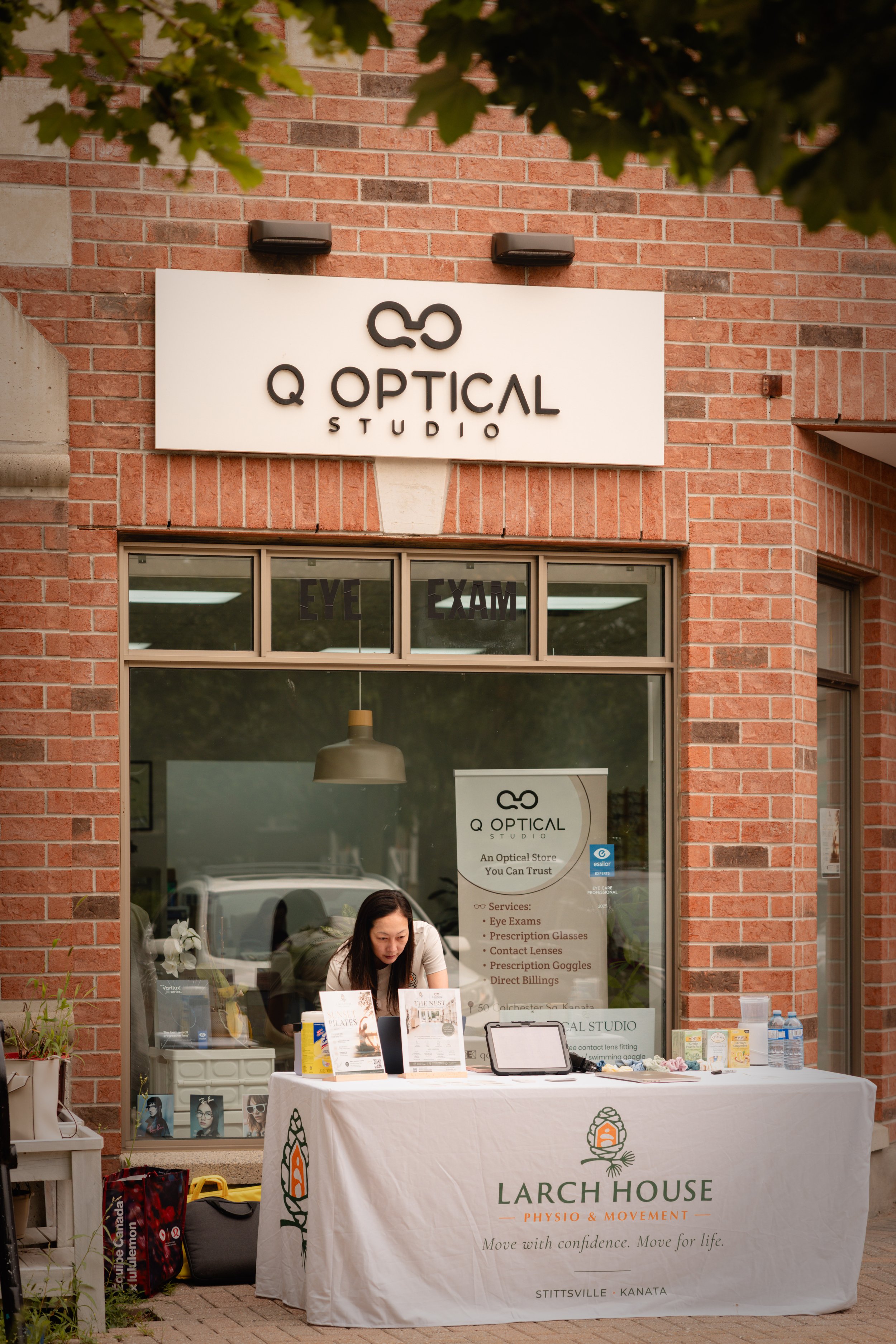 A woman sitting at a table outside a laser eye care center, with a sign reading 'Larch House Physio & Movement' and a list of services such as eye exams and prescription glasses. The table has brochures, a tablet, and bottles of water, under a large window of the brick building.