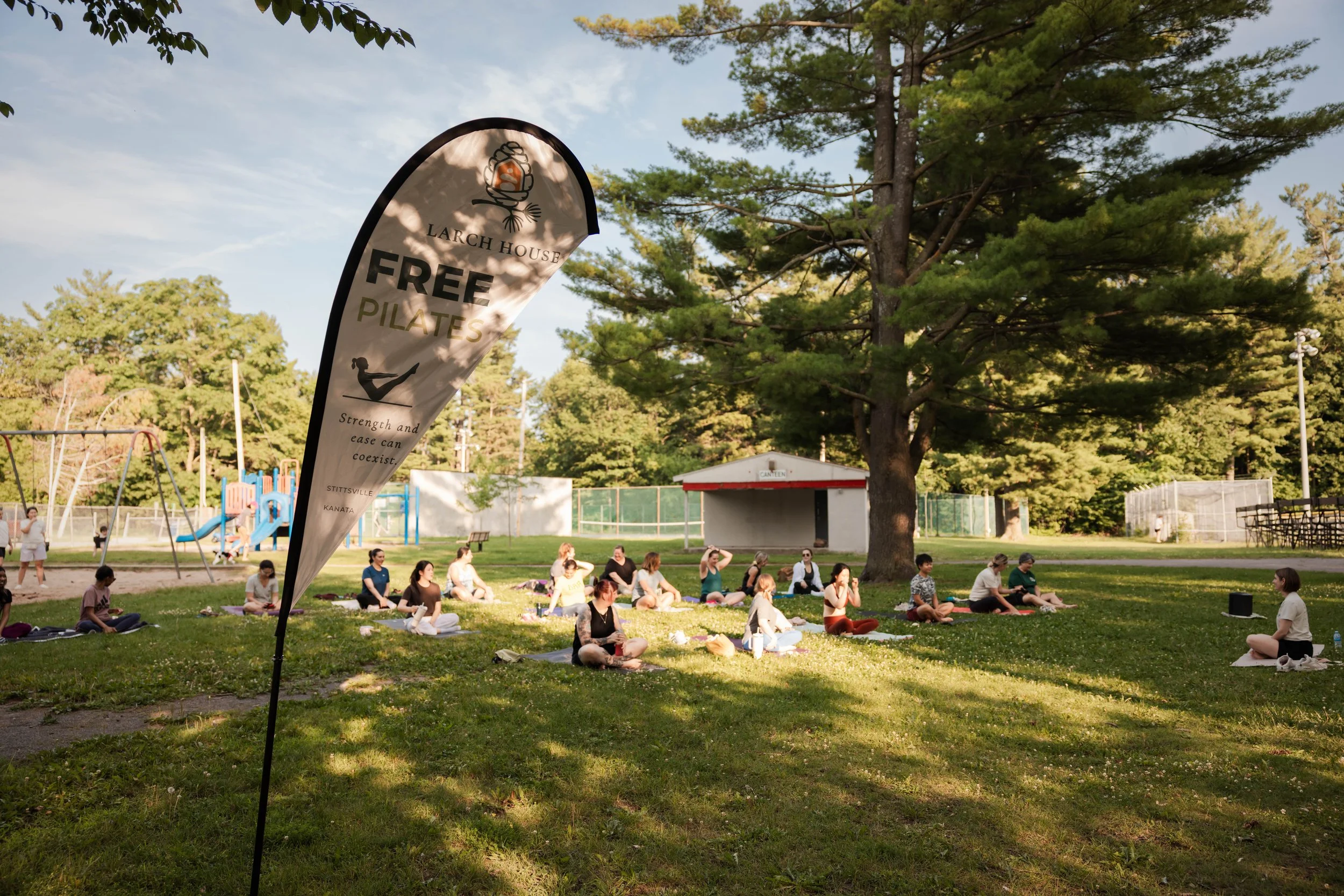 People practicing yoga outdoors in a park with a large tree, playground, and sports fields in the background, with a black and white banner advertising free Pilates classes.