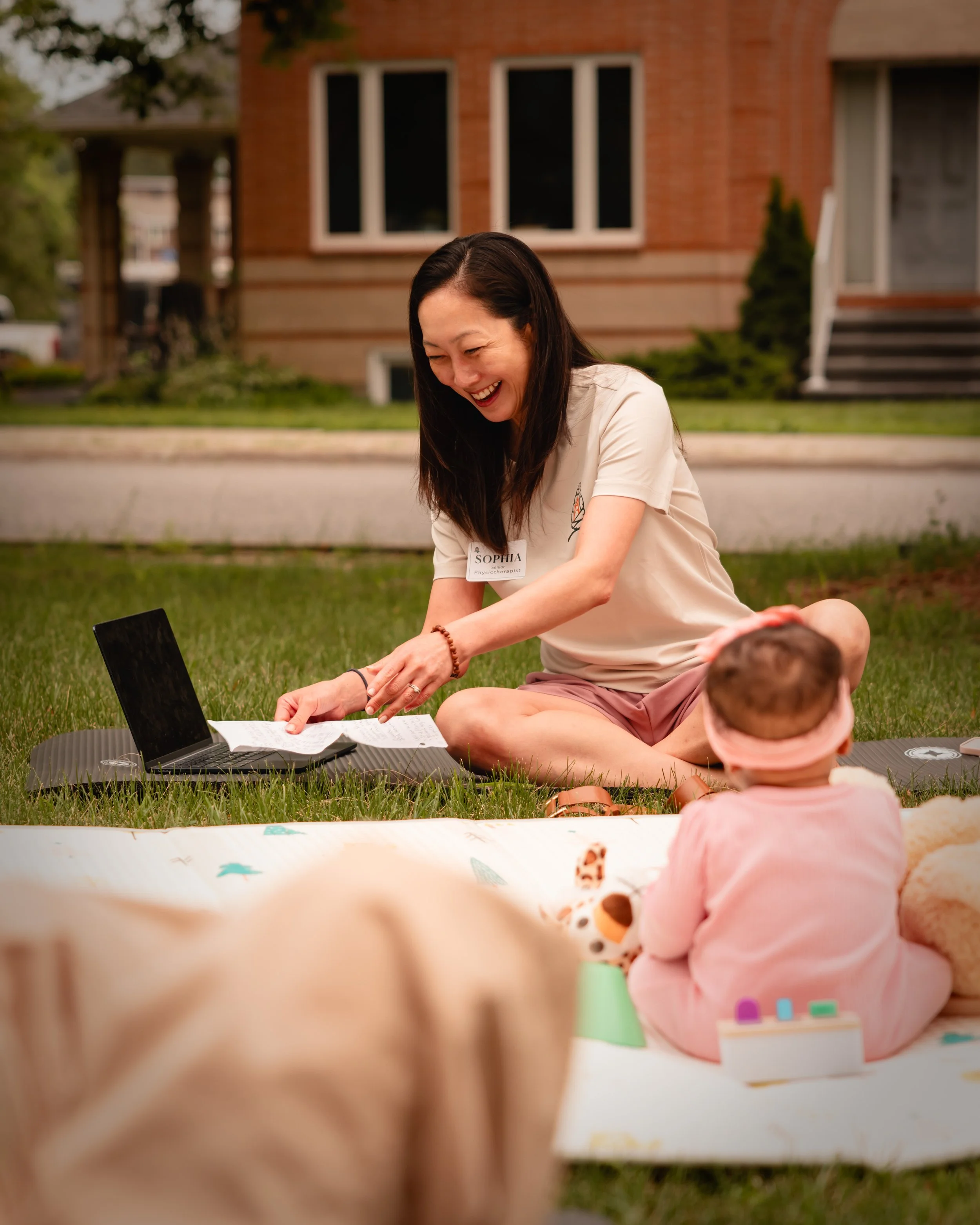 A woman with long dark hair, wearing a beige shirt with a name tag, is smiling and interacting with children during an outdoor activity on the grass in front of a brick house. The scene is lively and cheerful.