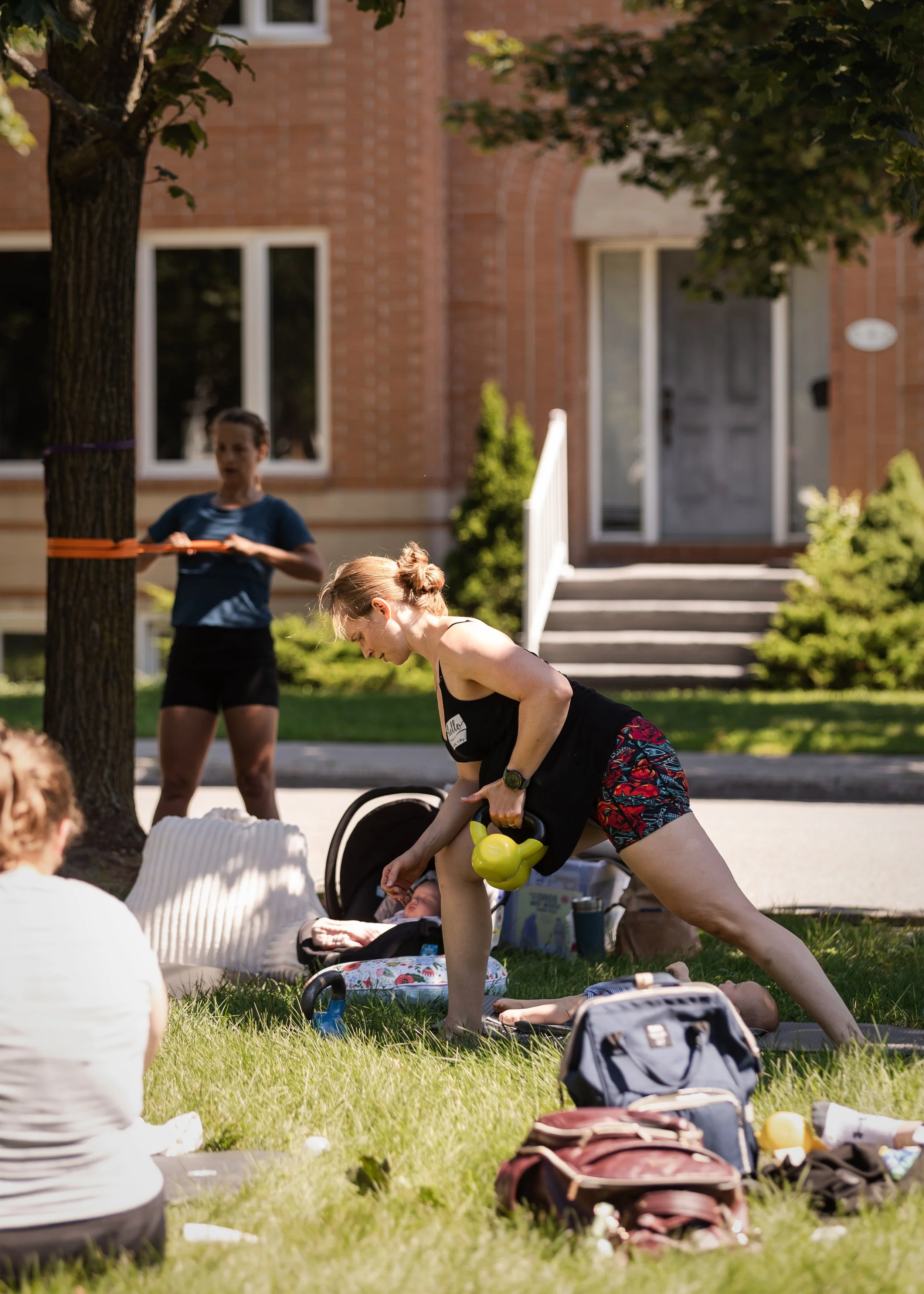 Two women performing outdoor exercises, one bending forward with a kettlebell and another standing behind her, in front of a brick building with steps and trees.