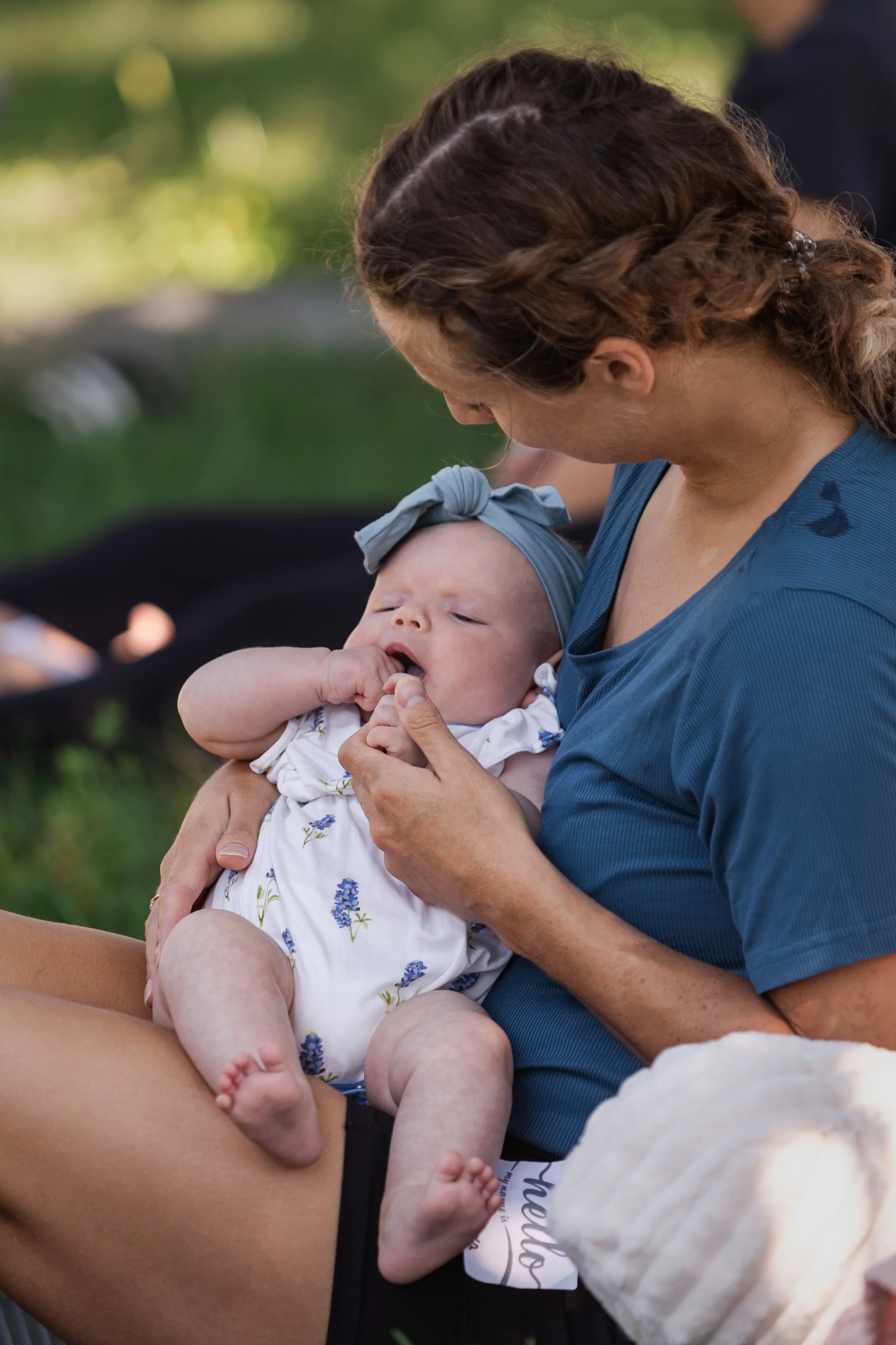 A woman with curly brown hair in a braid holds a baby with a gray headband and a white onesie with a floral pattern, sitting outdoors with a blurred green background.