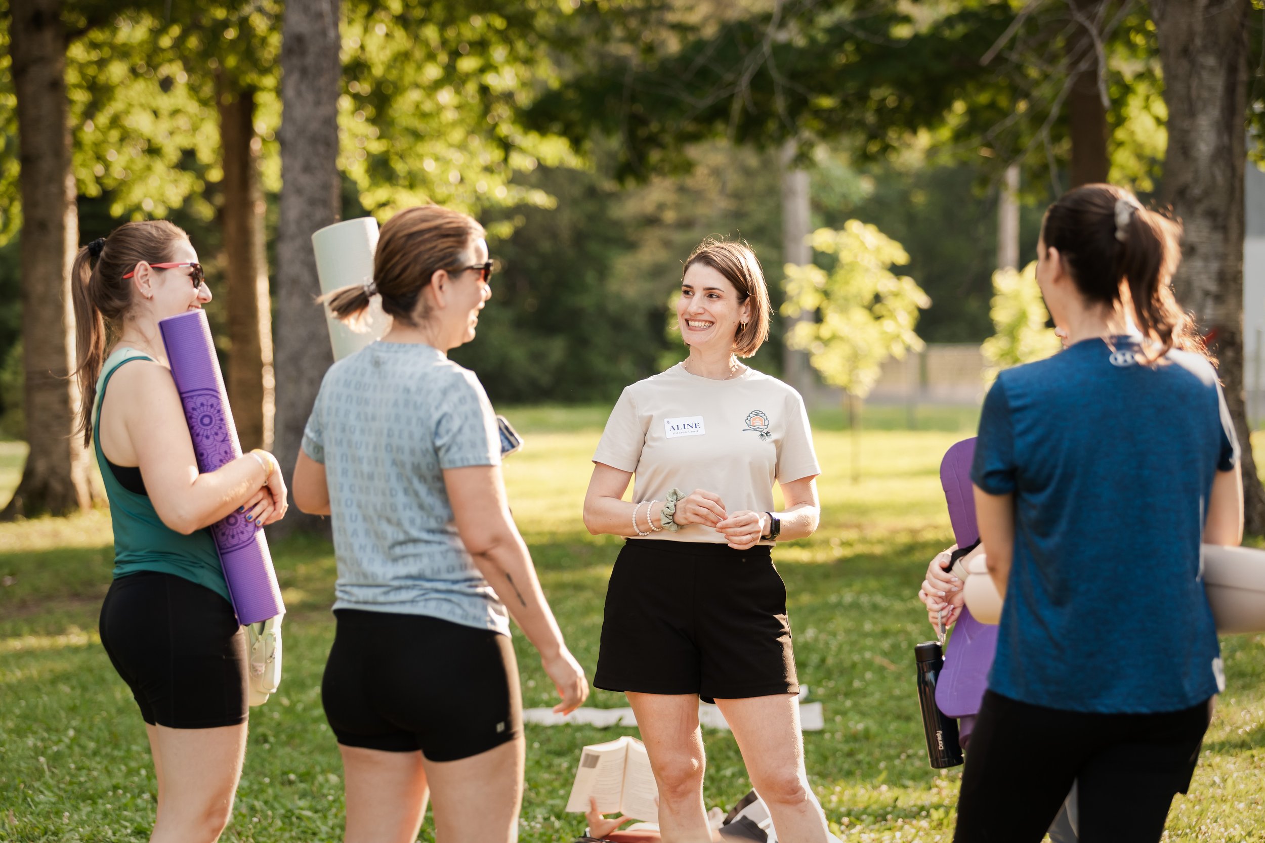 A group of women gathered outdoors in a park, engaging in conversation, with one woman smiling and wearing a white t-shirt, while others hold yoga mats and water bottles.
