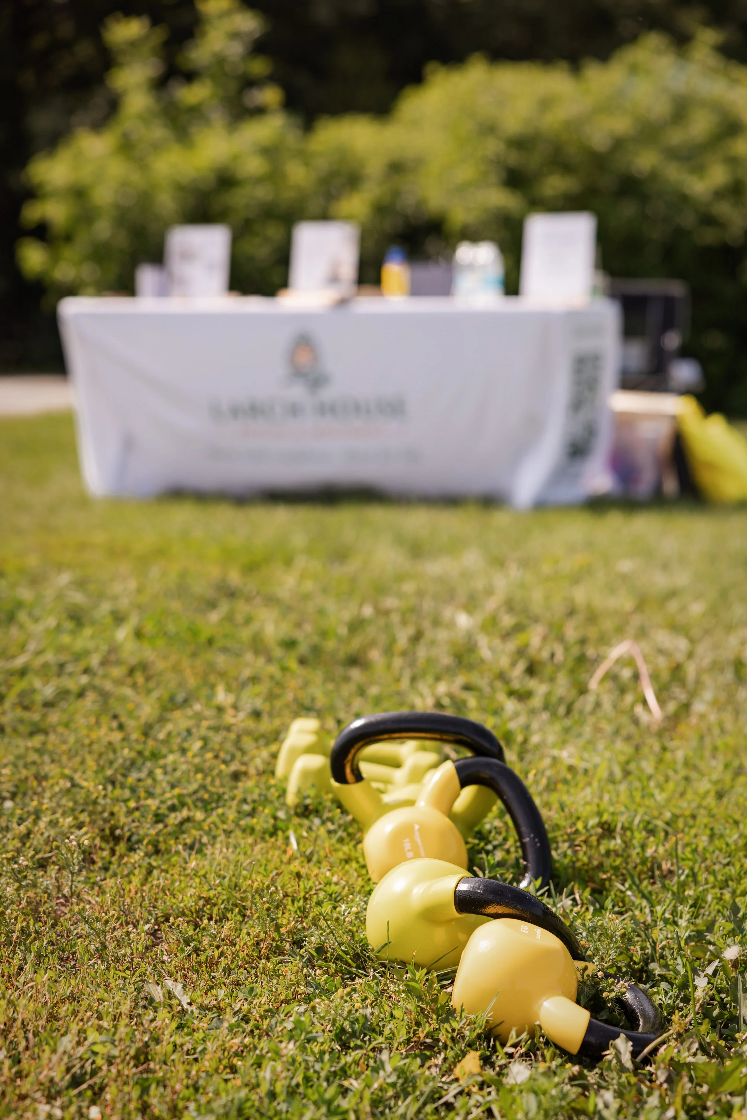 Kettlebells lined up on grass with yellow and black handles in the foreground, a blurred table with informational materials in the background surrounded by trees.