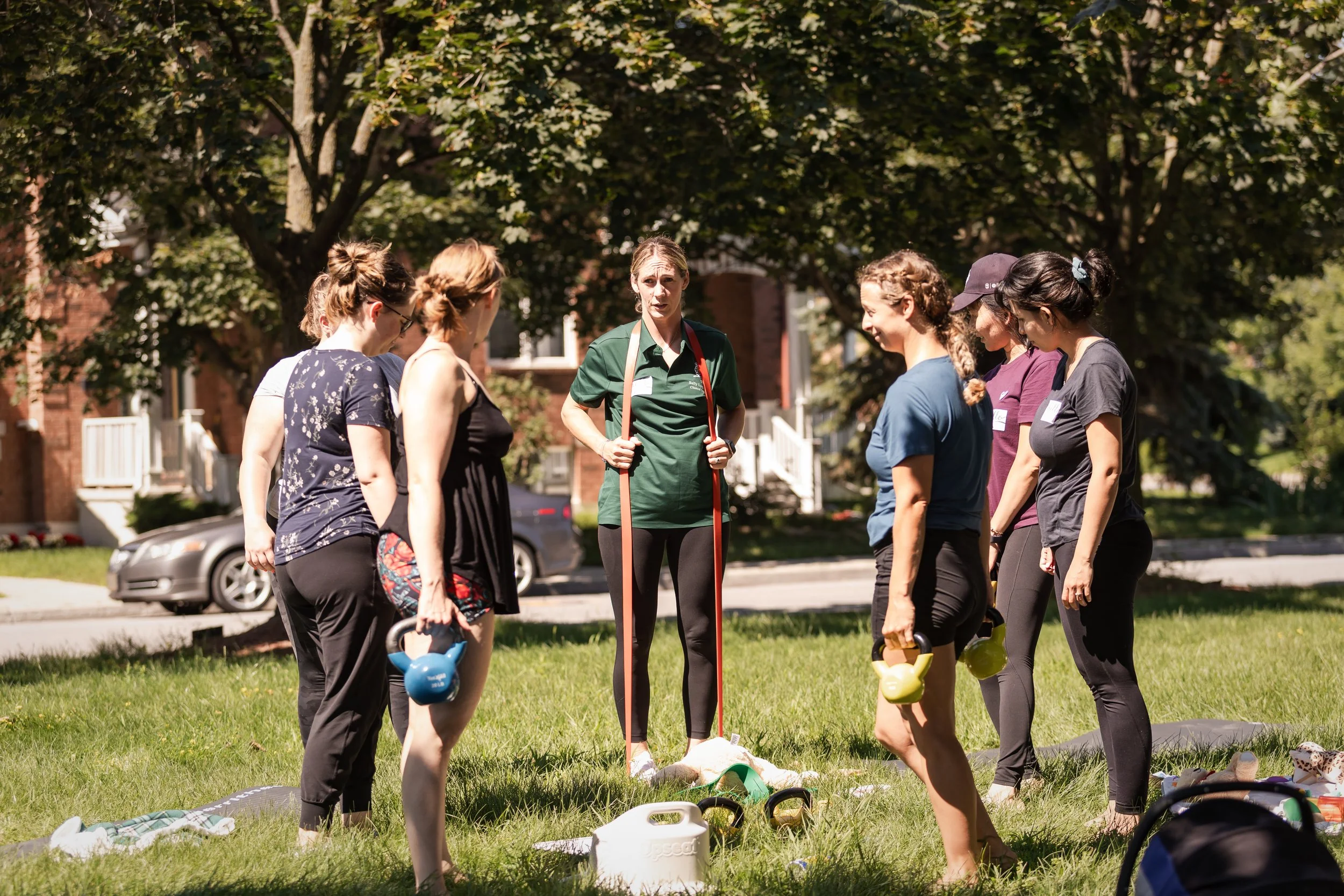A group of women participating in an outdoor fitness class under trees with a woman instructor demonstrating exercises.