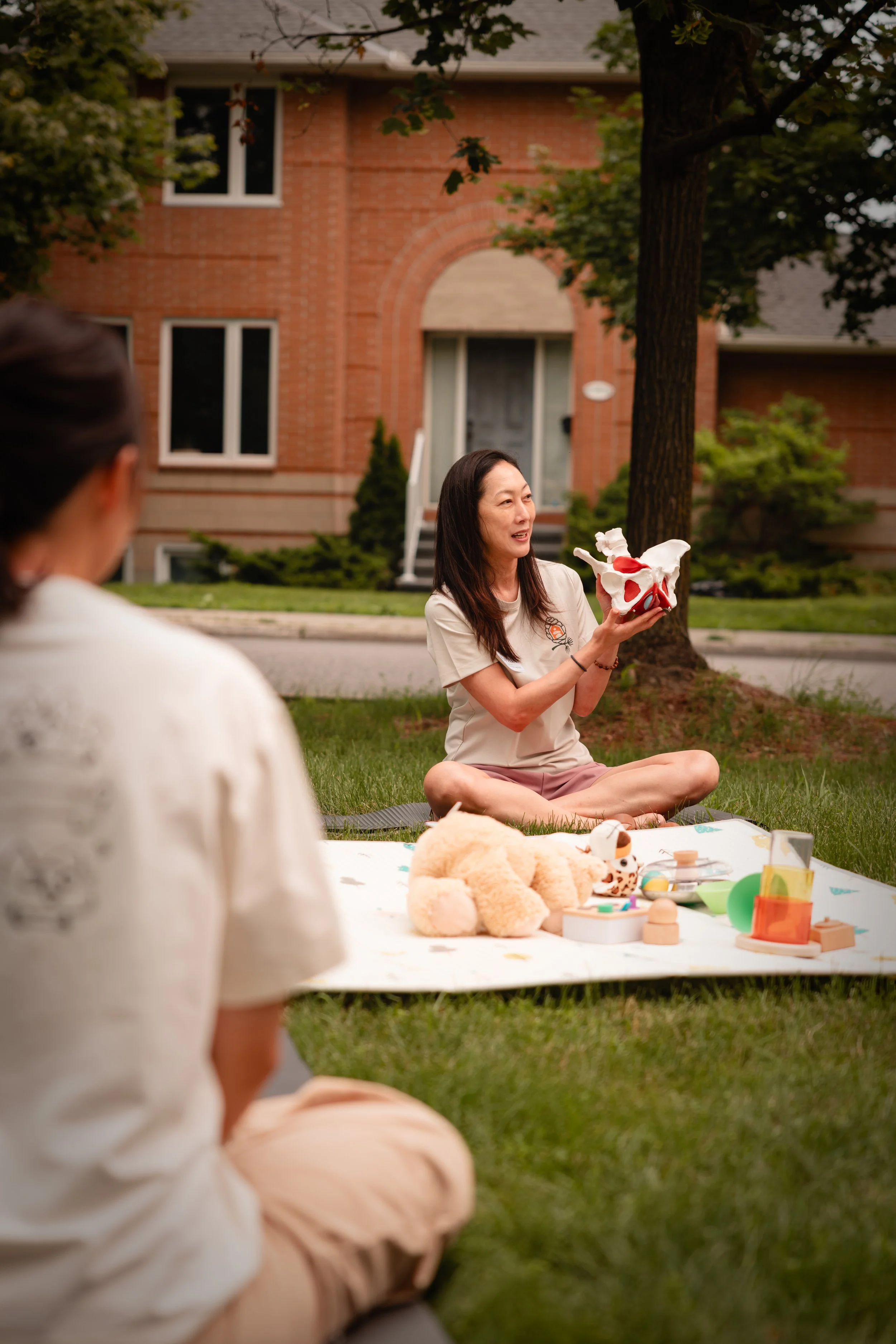 A woman sitting cross-legged on the grass holding a skull model, with children and toys on a blanket nearby. The scene is outside a brick house with trees.