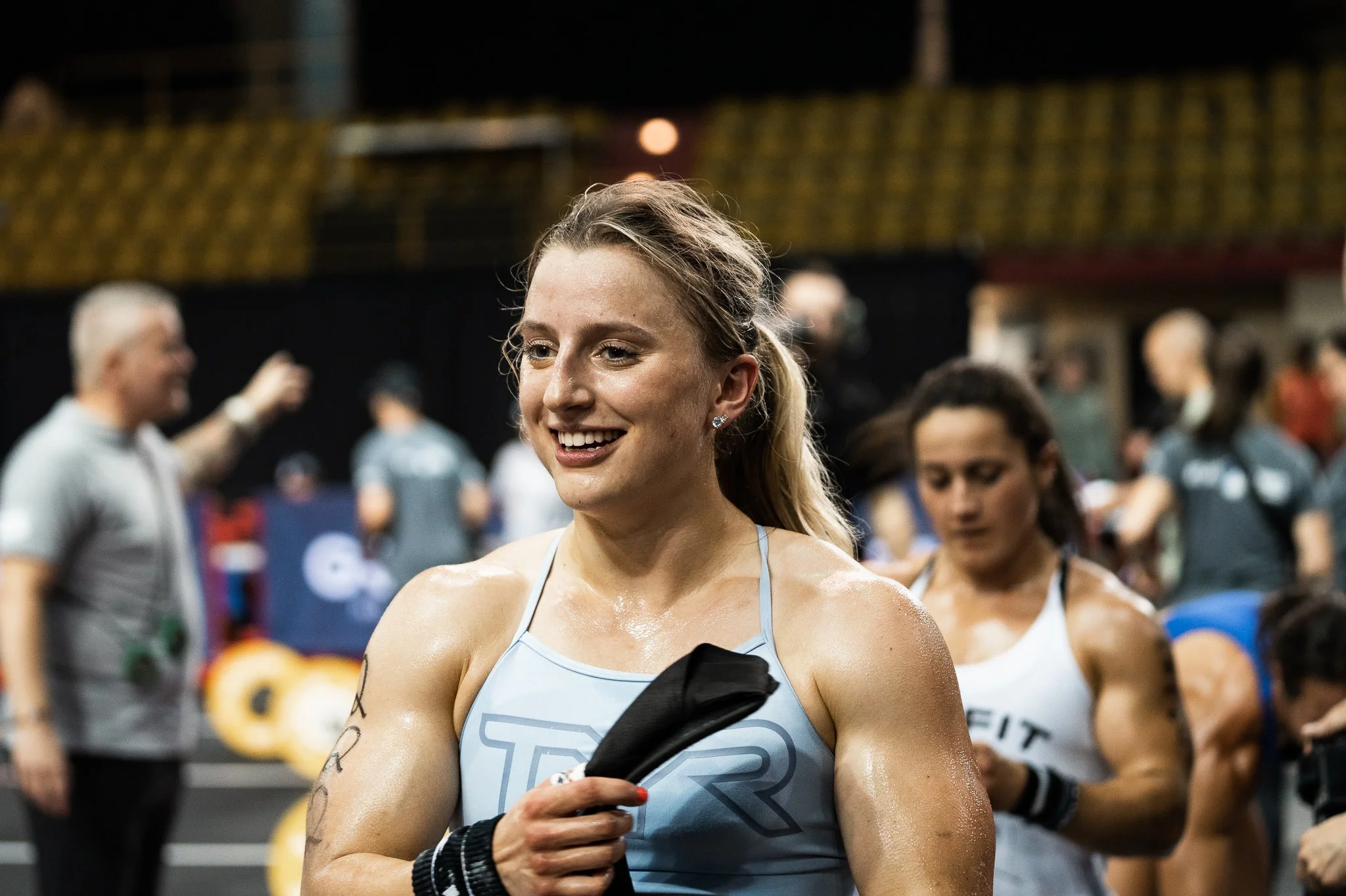 A smiling woman with blonde hair tied in a ponytail, wearing a tank top and black wristbands, holding a black object during a fitness event. Blurred people and gym equipment are visible in the background.