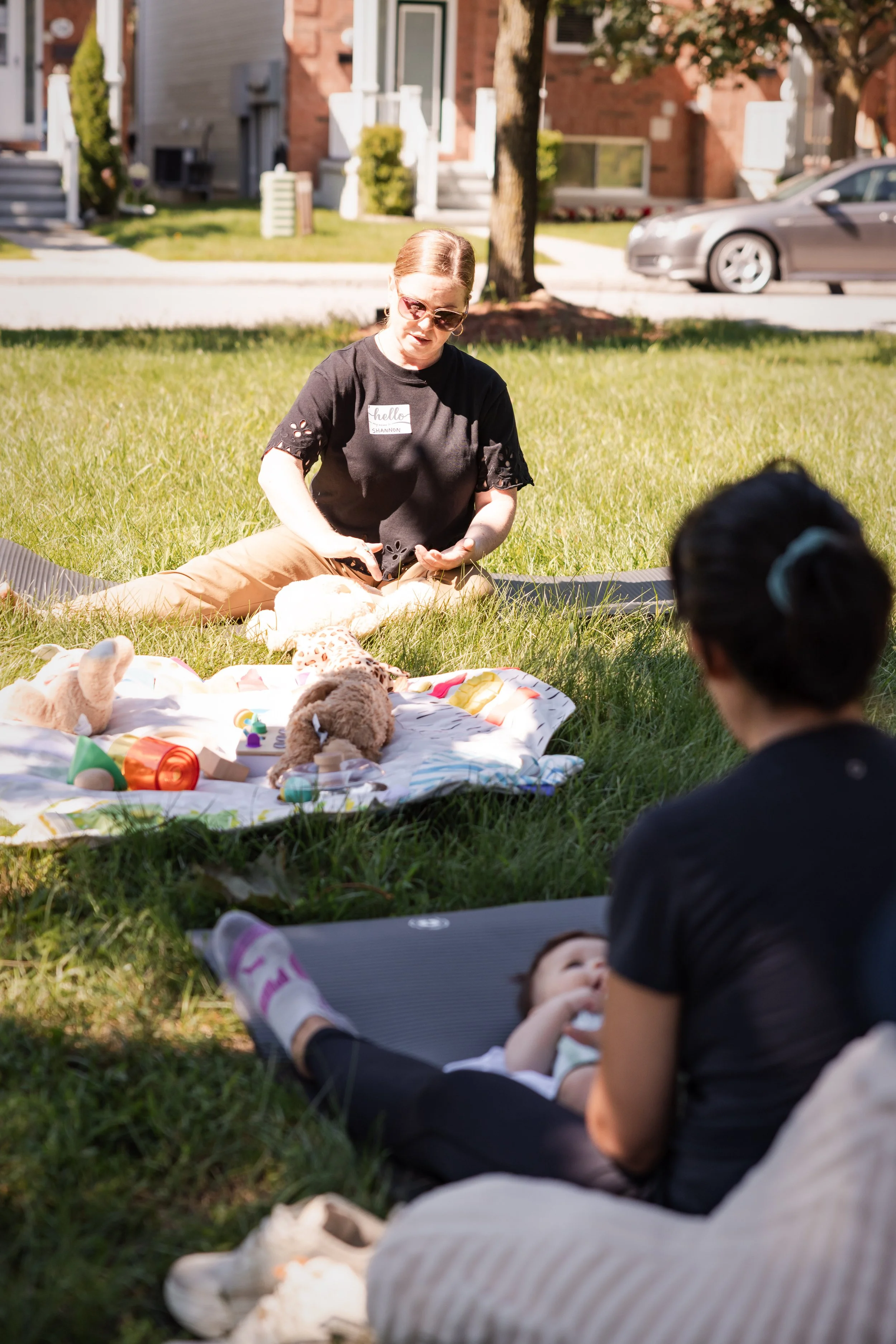 A woman with sunglasses, sitting on the grass during daytime, surrounded by children and stuffed animals, in a residential neighborhood.