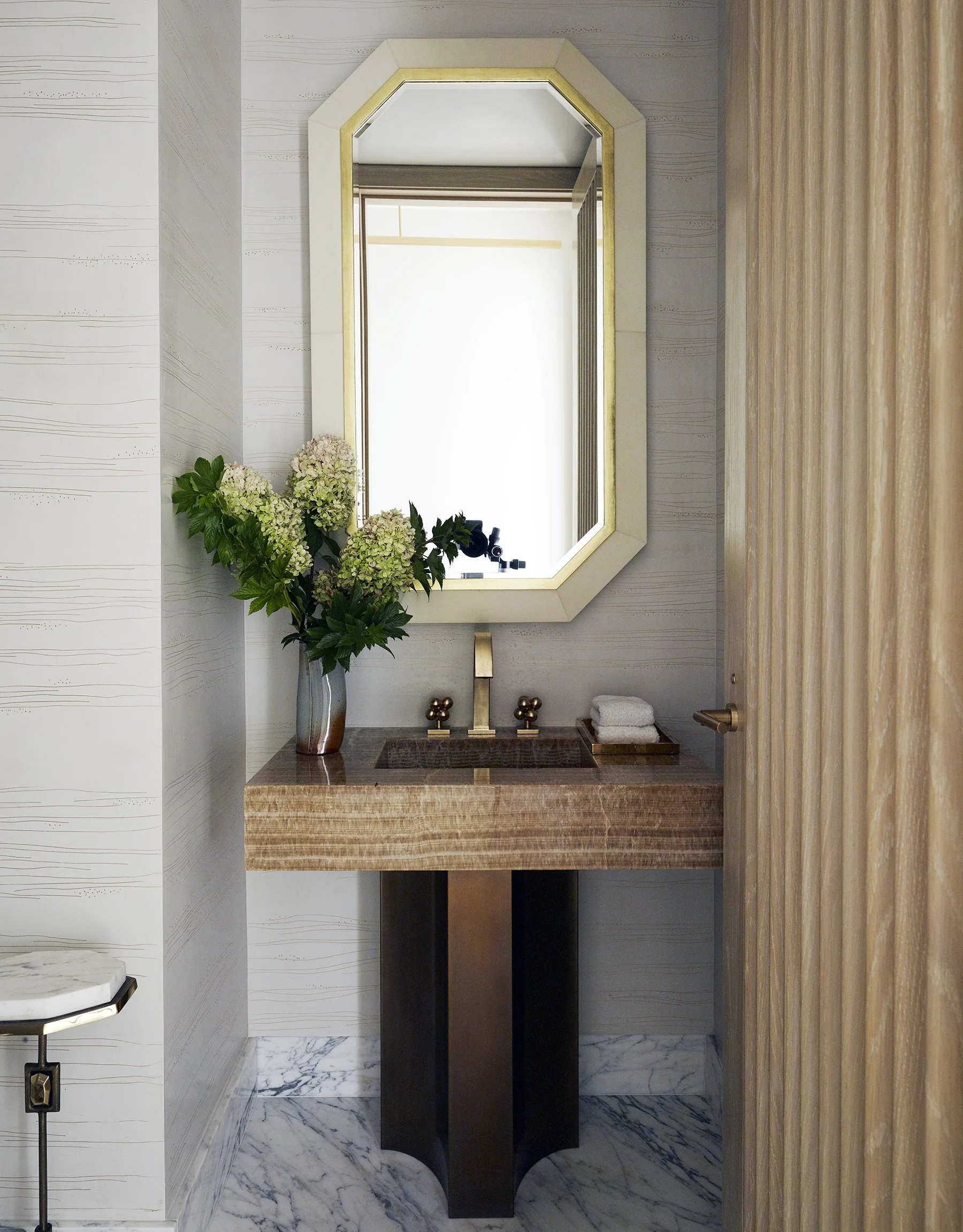 Elegant powder room with travertine floating vanity, brass fixtures, octagonal gold-framed mirror, textured grasscloth wallpaper, and Calacatta marble floor, 432 Park Avenue penthouse