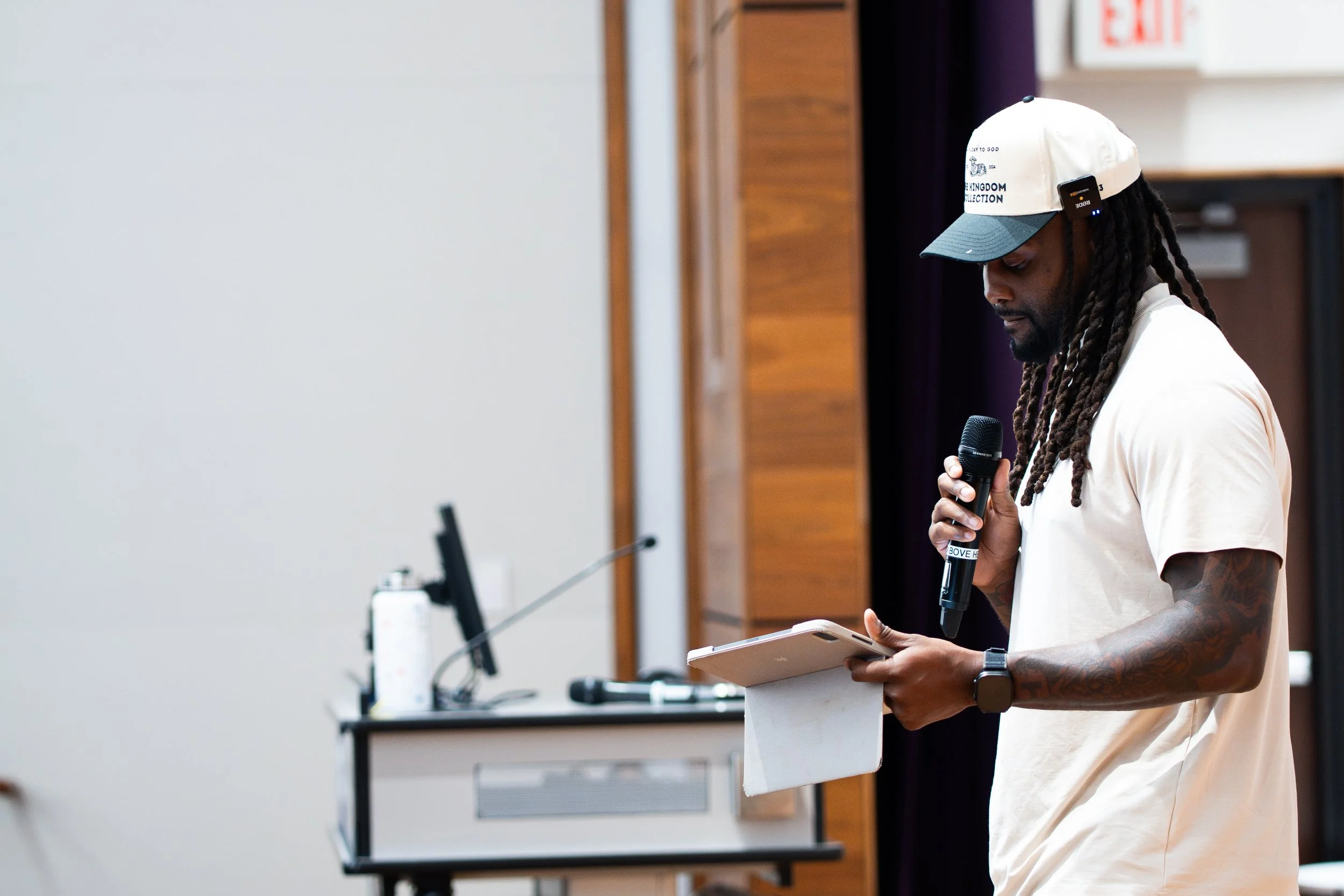 A man with long dreadlocks and tattoos on his right arm, wearing a white t-shirt, a cap, and a smartwatch, is holding a microphone in one hand and a tablet in the other, speaking at an indoor event.