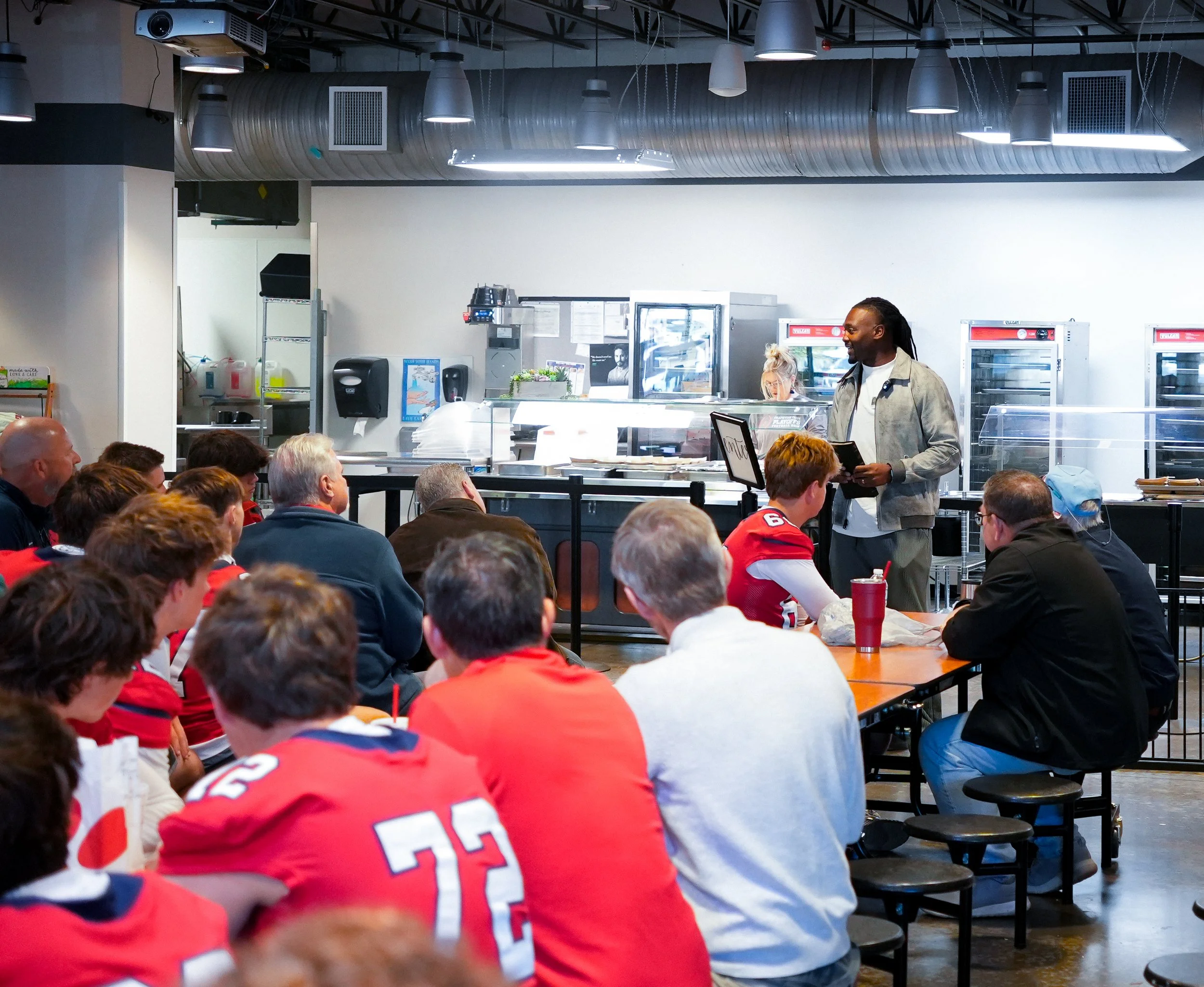 Athletes sitting at tables in a cafeteria or restaurant, with a man standing behind a counter and a woman wearing a headset in the background. The room has industrial ceiling with exposed ductwork and pendant lights.