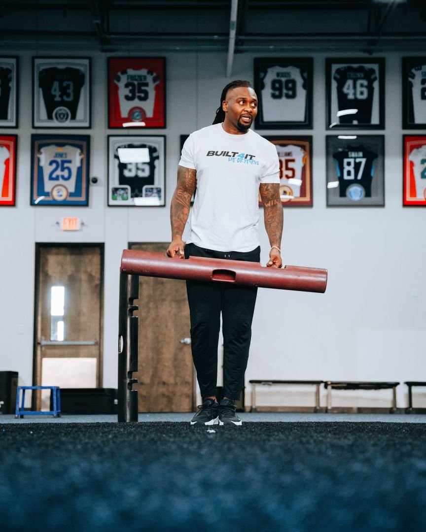 Kavon Frazier holding a padded training dummy in a gym with framed NFL and professional jerseys on the wall behind him.