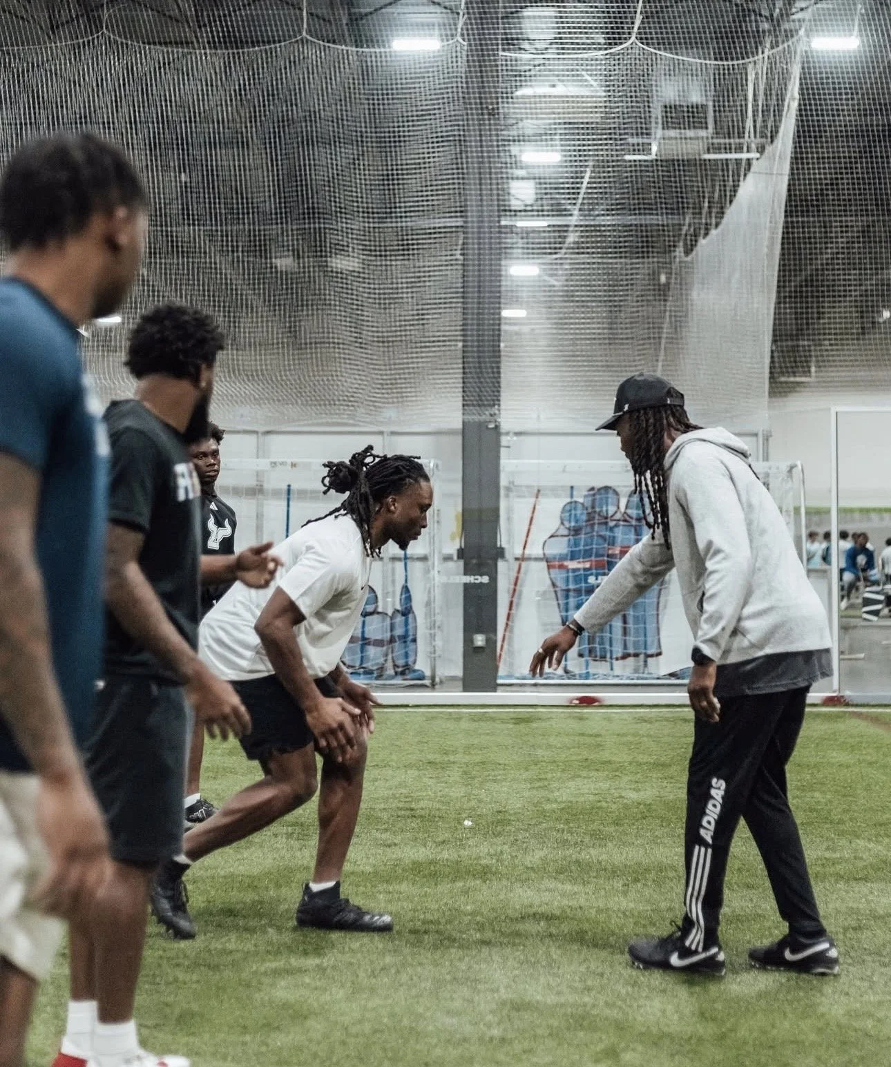A group of athletes participating in a training session on an indoor field, some of them are in a defensive stance while the coach or trainer is giving instructions, with sports equipment and netting in the background.