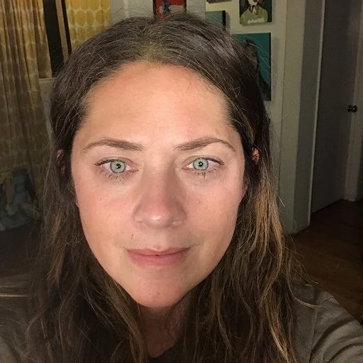 A woman with long, wavy brown hair and green eyes taking a selfie indoors, with artwork on the wall behind her.