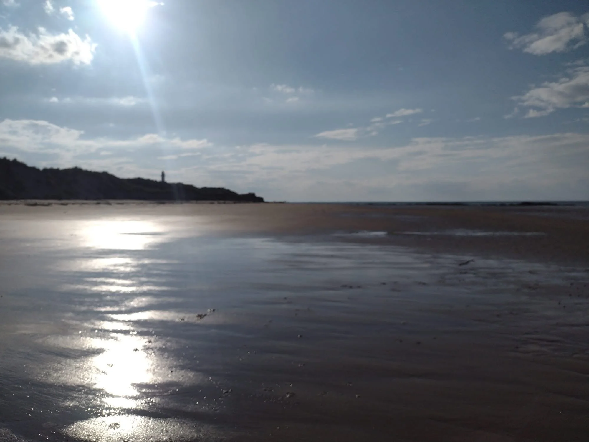 A beach scene with the sun shining brightly in a partly cloudy sky. The shoreline with wet sand reflecting the sunlight is visible, and a distant headland with a solitary figure standing on it is seen against the horizon.