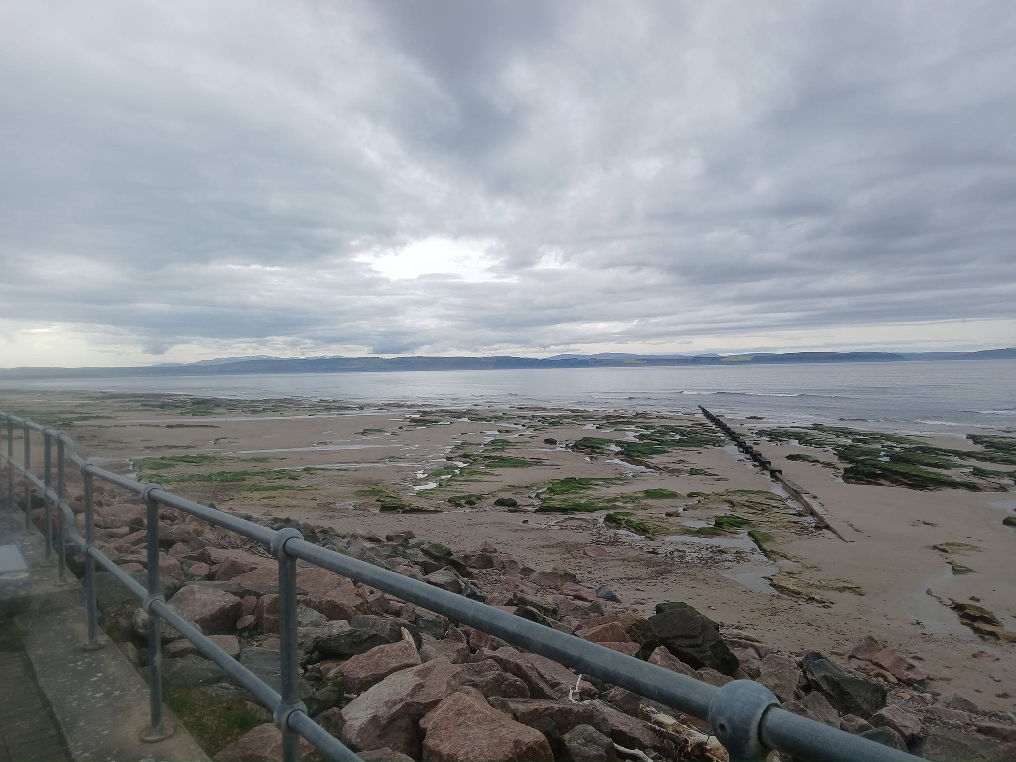 Overcast sky over a coastal area with rocky shoreline, beach, and water, viewed from a promenade with a metal railing.