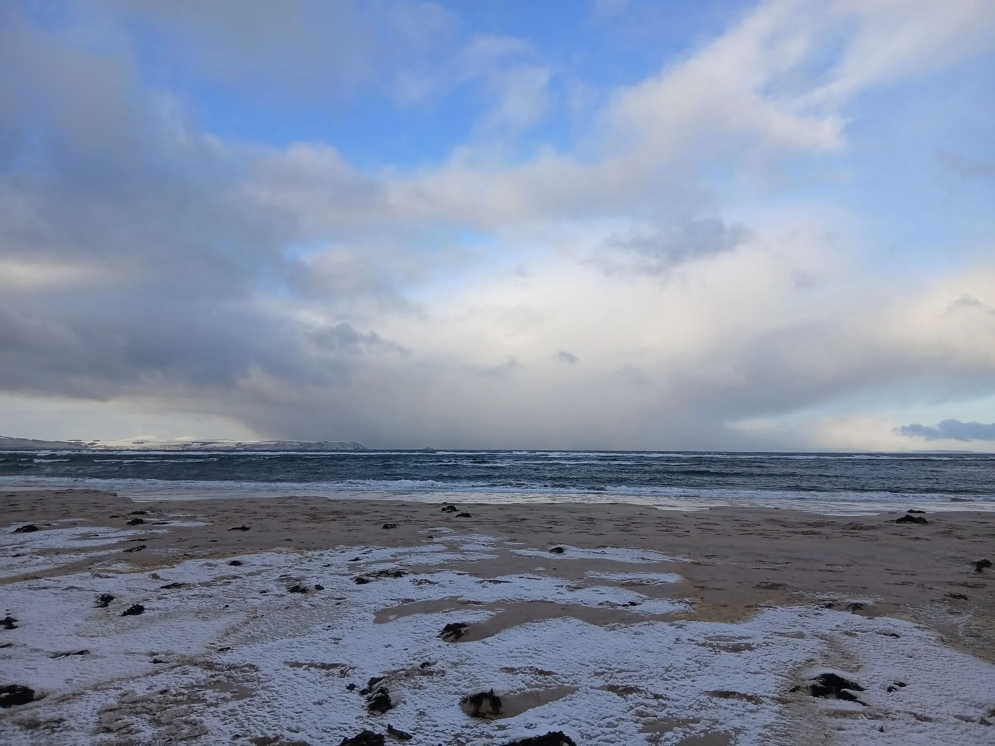 A beach scene with snow patches, dark wet sand, and distant shoreline under a partly cloudy sky.