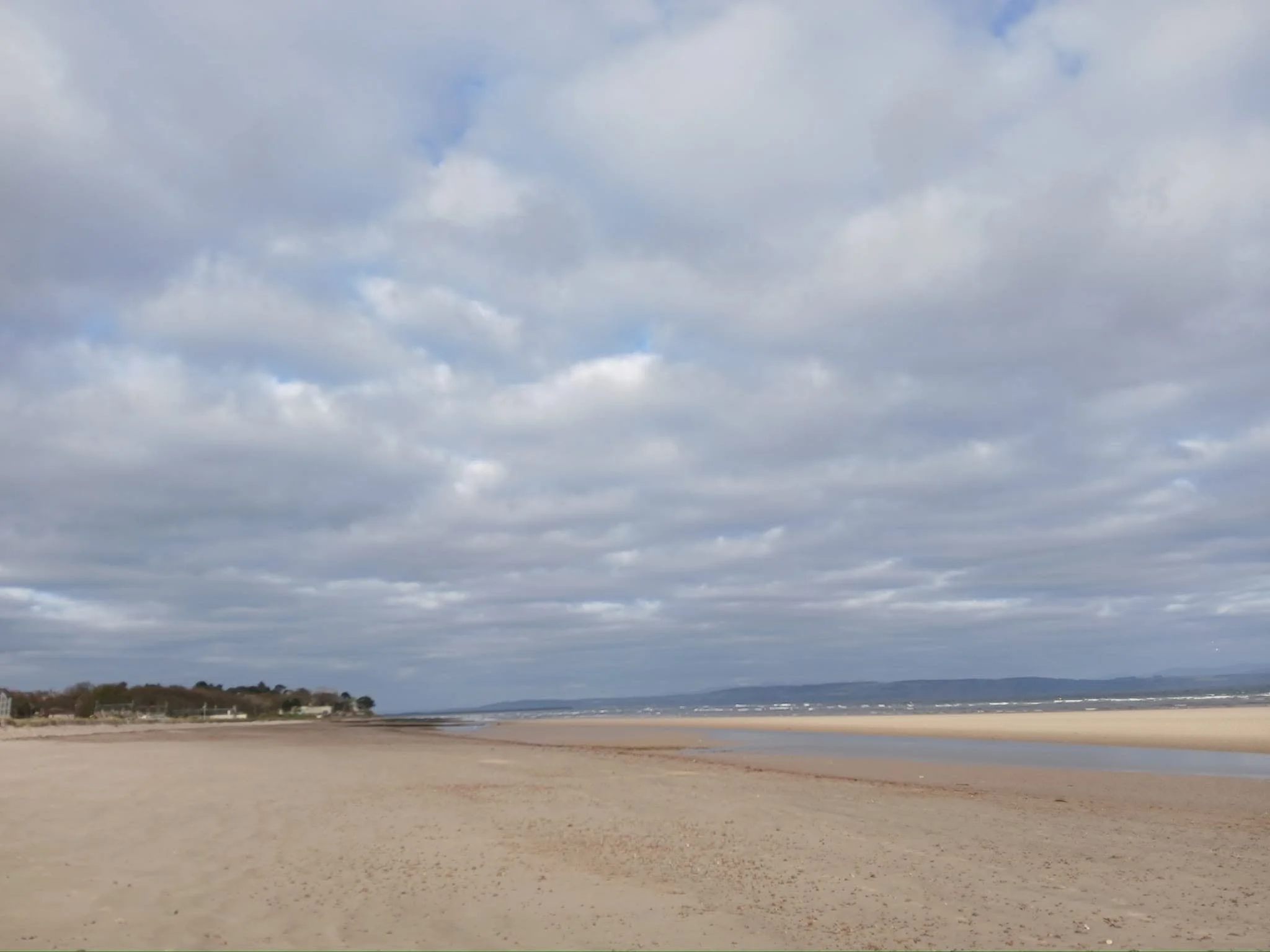 A wide beach with smooth sand, a few scattered trees, and distant hills under a cloudy sky.