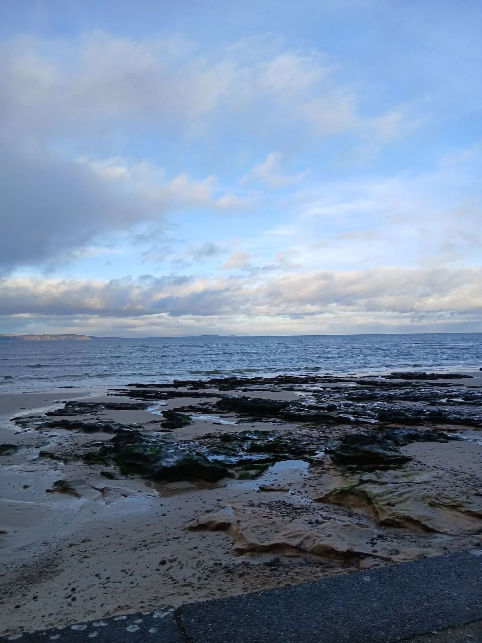 Cloudy sky over a calm ocean with rocks and sand in the foreground.