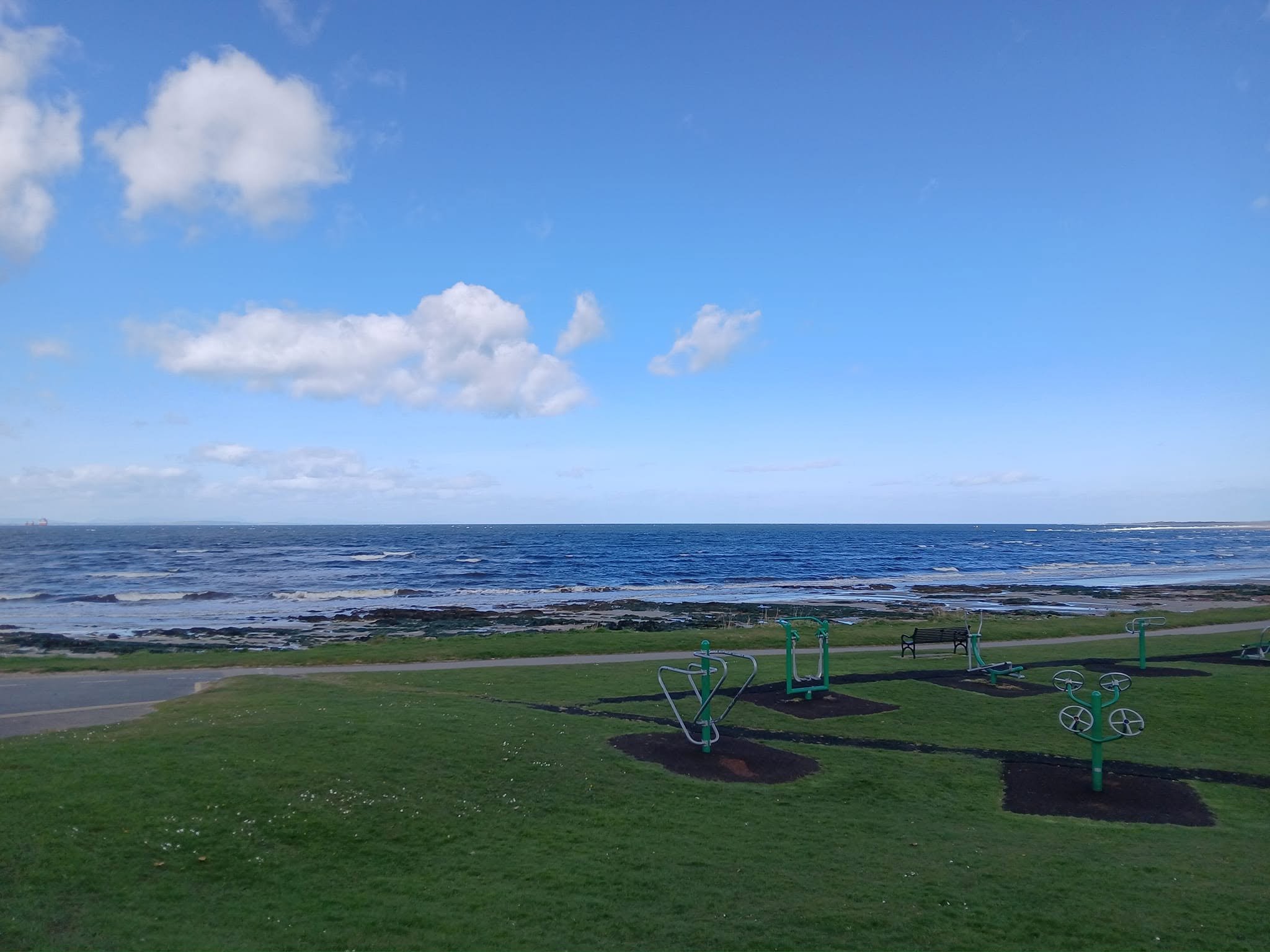 A park by the ocean with a grassy area, benches, outdoor exercise equipment, and a view of the water and sky.