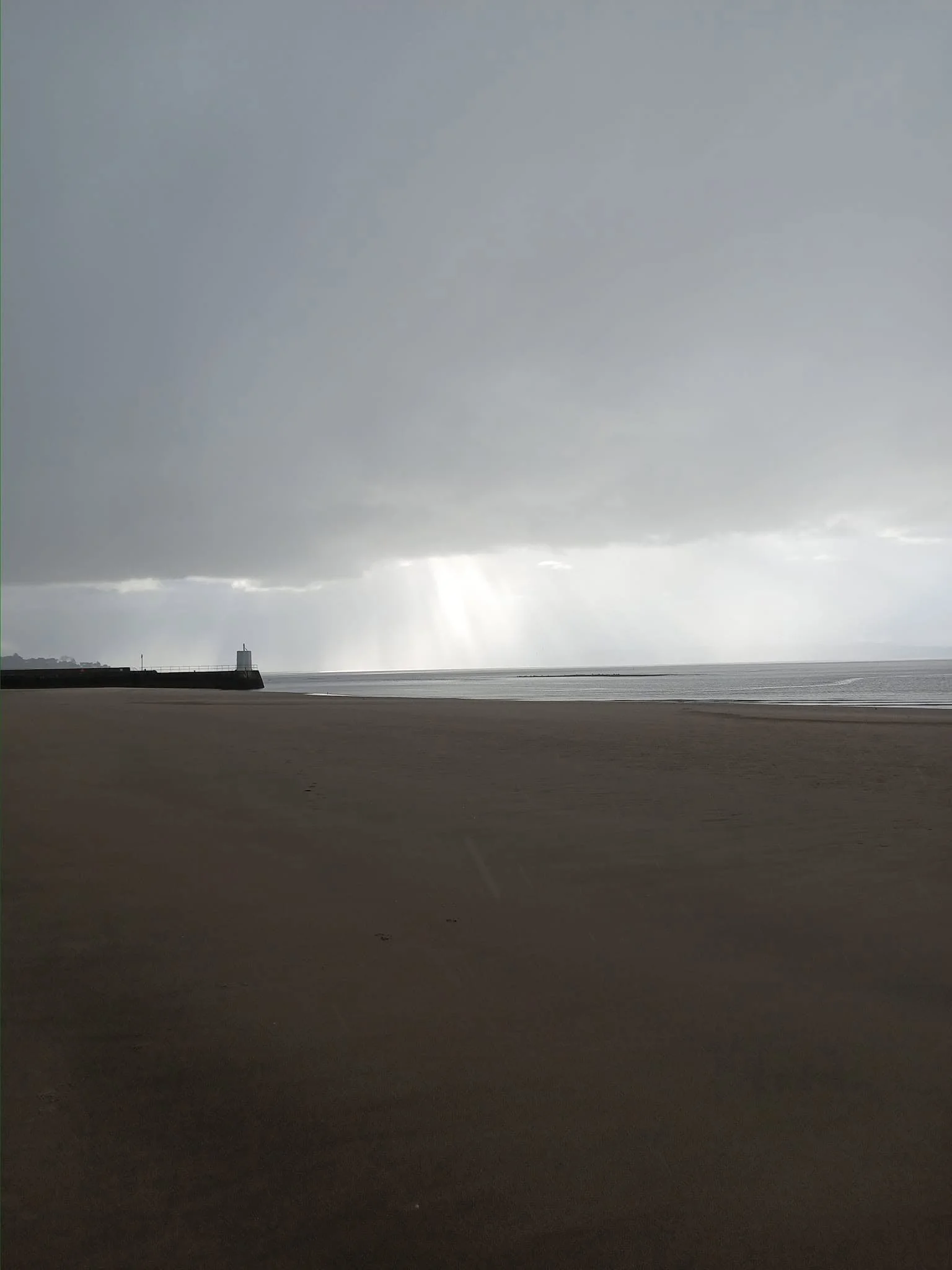 A beach with a structure near the water, a cloudy sky, and rays of sunlight breaking through the clouds over the ocean.